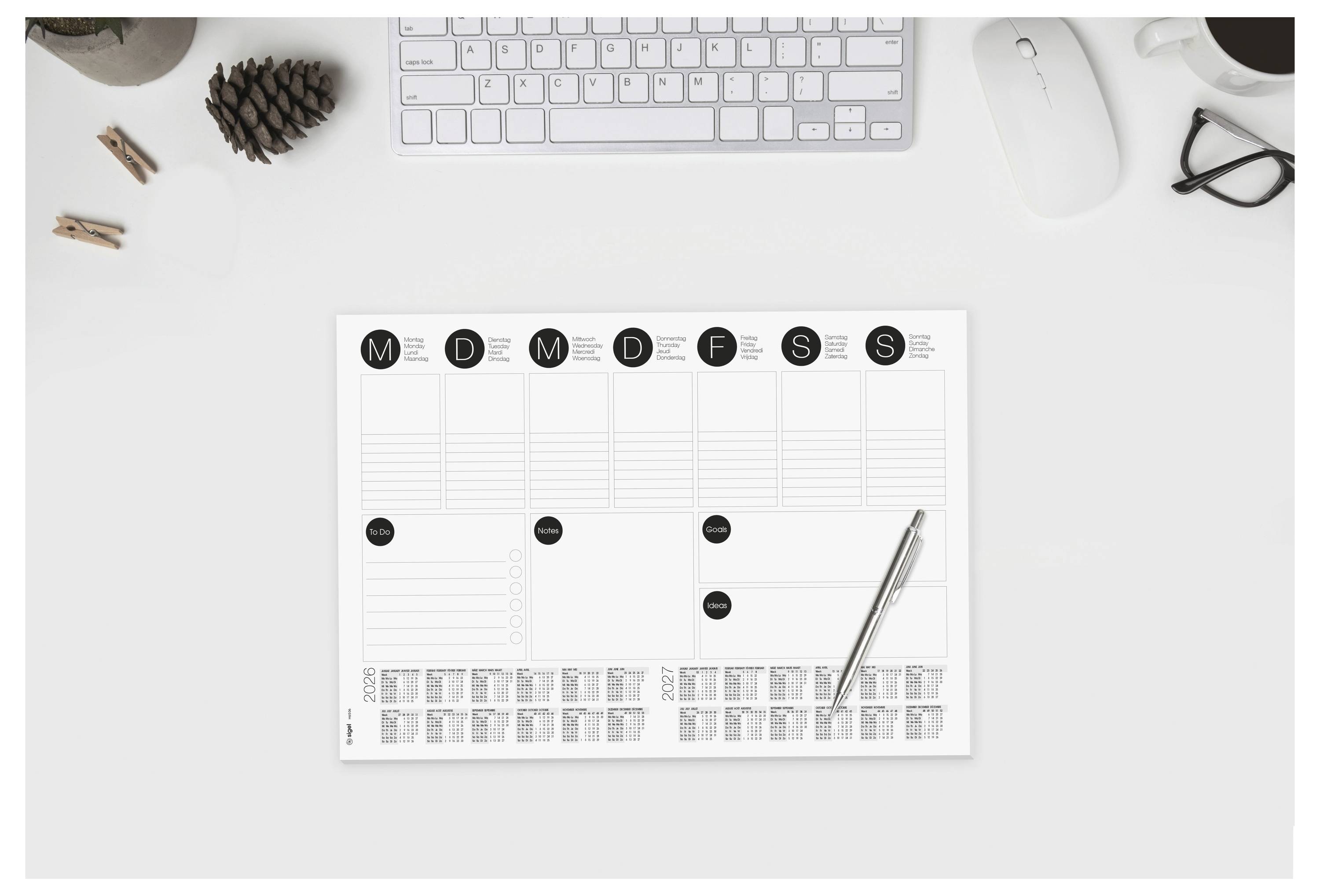 Minimalistischer Schreibtisch mit einem Wochenplaner mit den Beschriftungen M, D, M, D, F, S, S, einem diagonal darauf gelegten Stift und Büroartikeln drumherum.