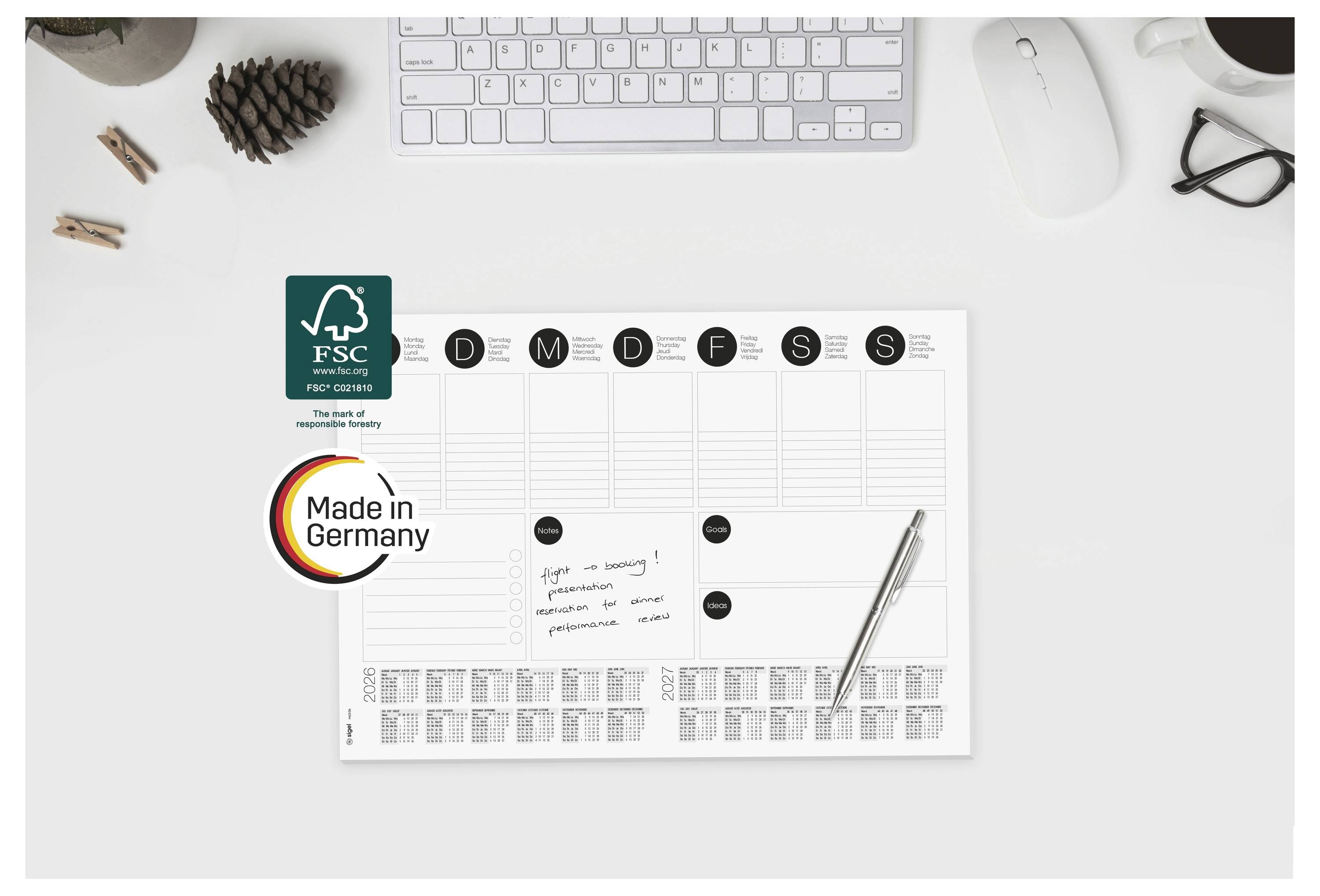 Draufsicht eines Schreibtischplaners mit FSC- und „Made in Germany"-Etiketten, einem Stift, einer Tastatur, einer Maus und Bürozubehör auf einem weißen Arbeitsplatz.