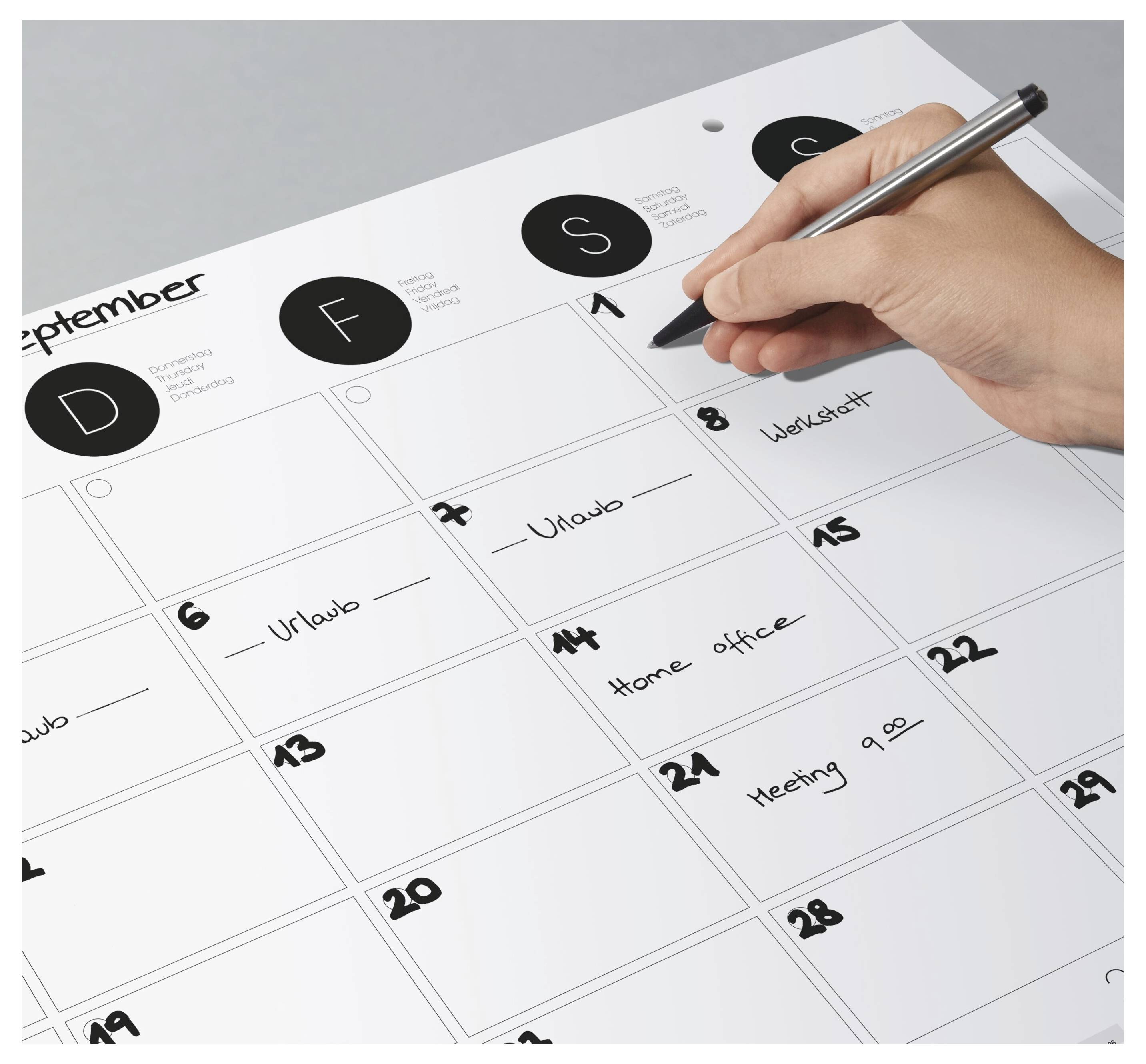 Eine Hand schreibt einen Termin in einen Septemberkalender, wobei mehrere Daten in Deutsch beschriftet sind, wie z.B. 'Urlaub', 'Werkstatt', 'Homeoffice' und 'Meeting 9.00'.