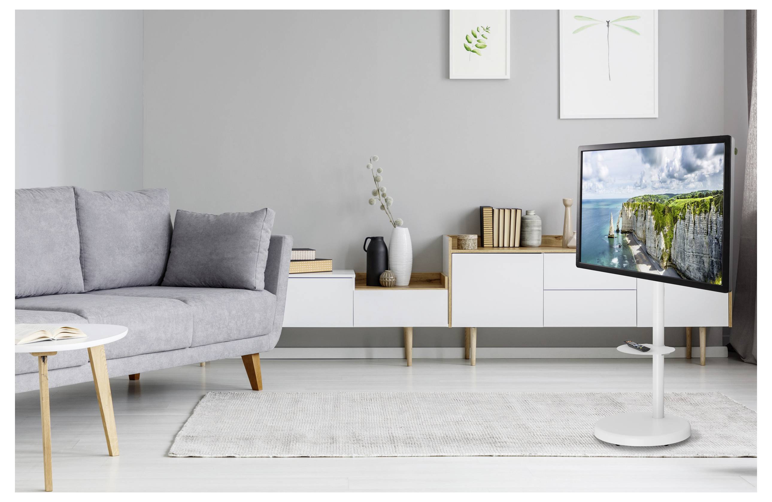 Ein moderner Wohnraum mit einem grauen Sofa, weißem Couchtisch, an der Wand montiertem Fernseher, der eine malerische Küstenszene zeigt, und minimalistischer Dekoration.