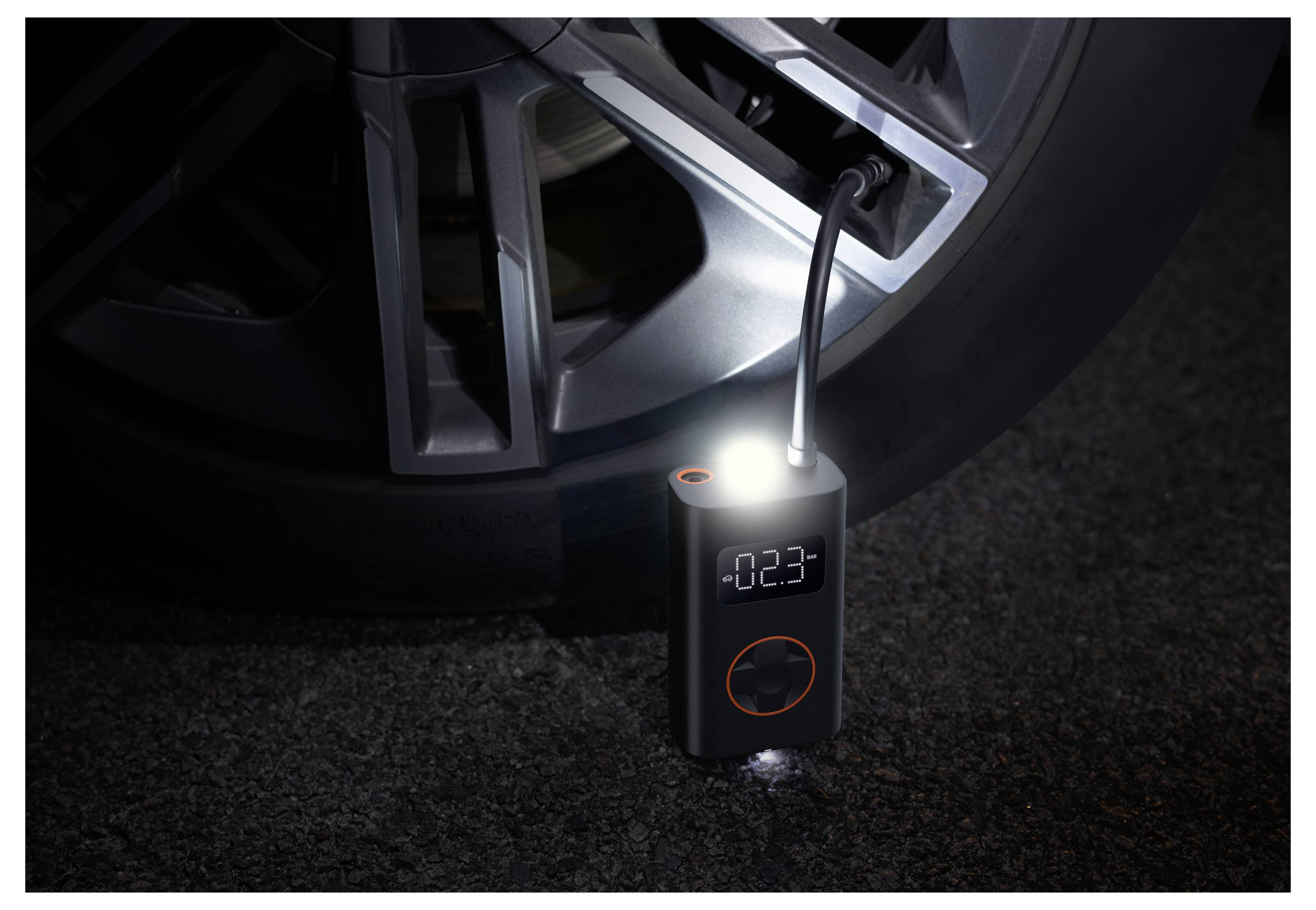 Ein tragbarer elektronischer Reifenaufpumper ist an einem Autoreifen angeschlossen und zeigt auf seinem Display den Luftdruck '0,23' an, beleuchtet von einem eingebauten Licht.