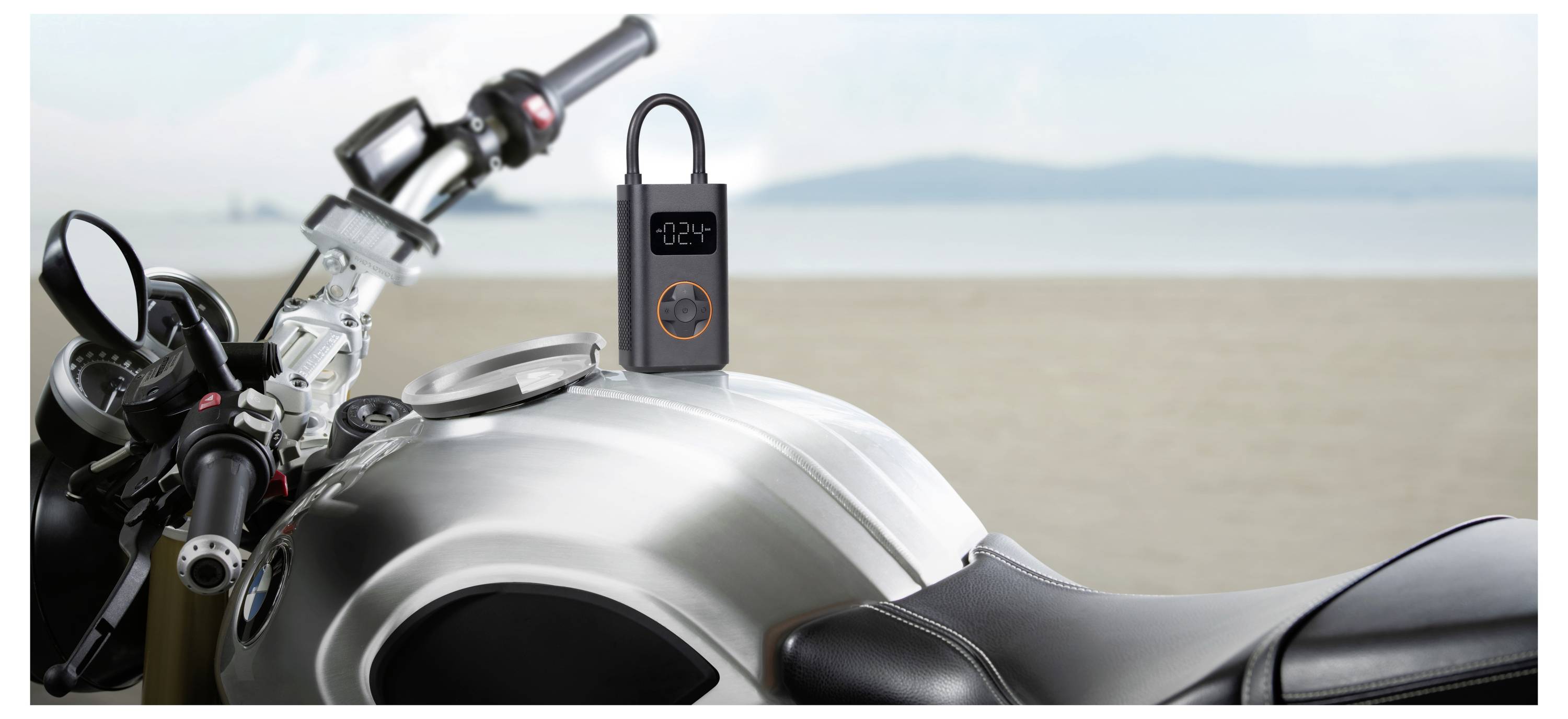 Eine tragbare Luftpumpe mit digitalem Display sitzt auf dem Kraftstofftank eines Motorrads am Strand.