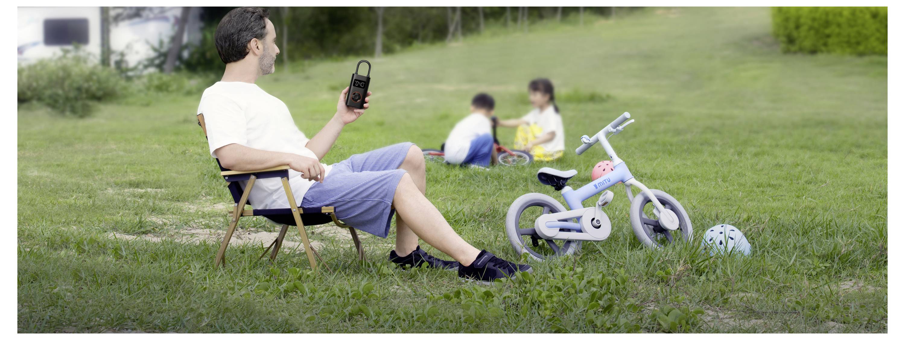 Ein Mann sitzt auf einem Stuhl im Park und schaut auf ein Smartphone, während zwei Kinder, die unscharf im Hintergrund sind, auf dem Gras sitzen, mit einem Fahrrad und Helmen in der Nähe.