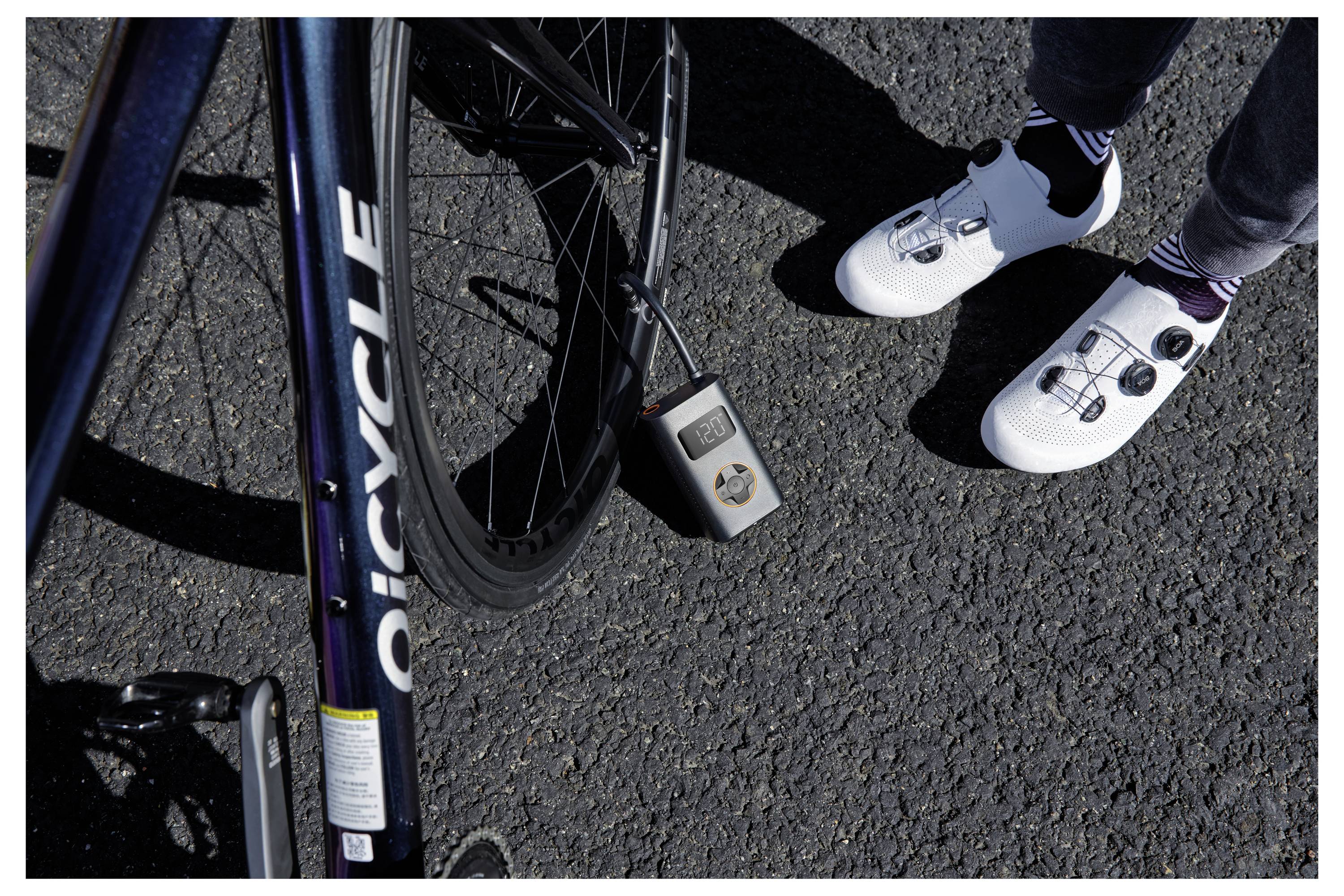 Ein schwarzes Fahrradrad, das mit einer digitalen Pumpe aufgepumpt wird, zeigt '002 PSI', neben einer Person mit weißen Radsportschuhen auf Asphalt.