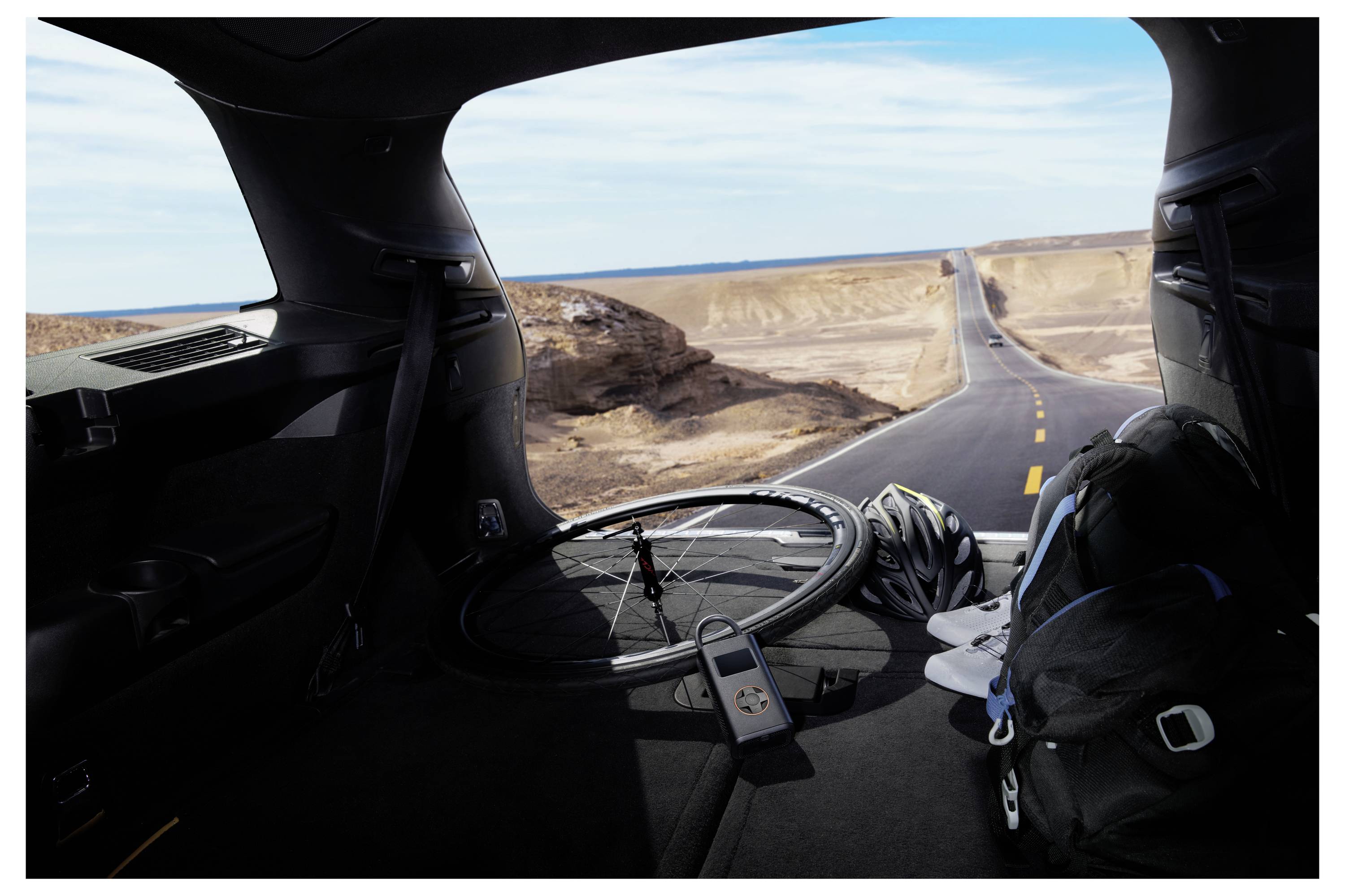 Blick von der Rückseite eines Autos, der ein Fahrradrad, einen Helm und Ausrüstung vor einer Straße zeigt, die sich in die Wüstenlandschaft unter einem blauen Himmel erstreckt.