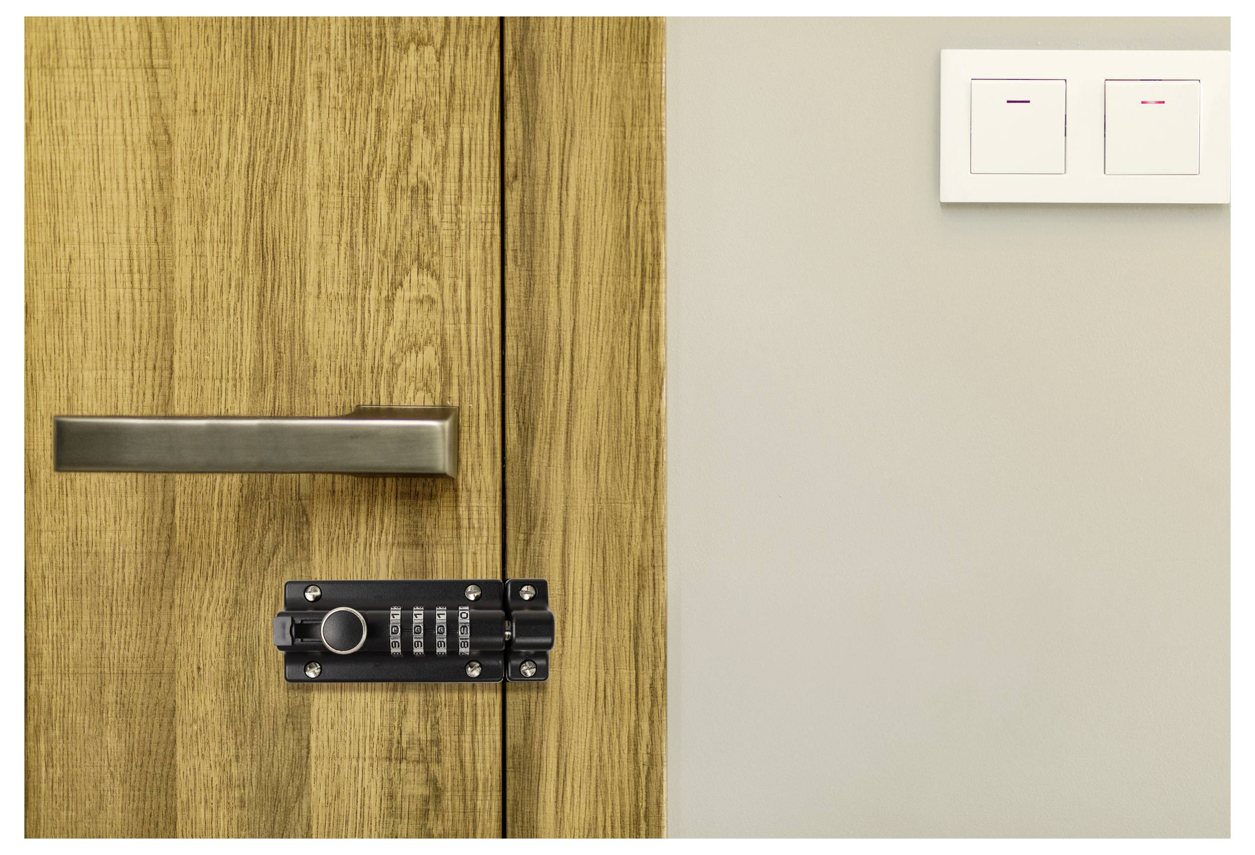 Eine verschlossene Holztür mit einem Griff und einem numerischen Tastenschloss neben einem Doppellichtschalter an einer beigen Wand.