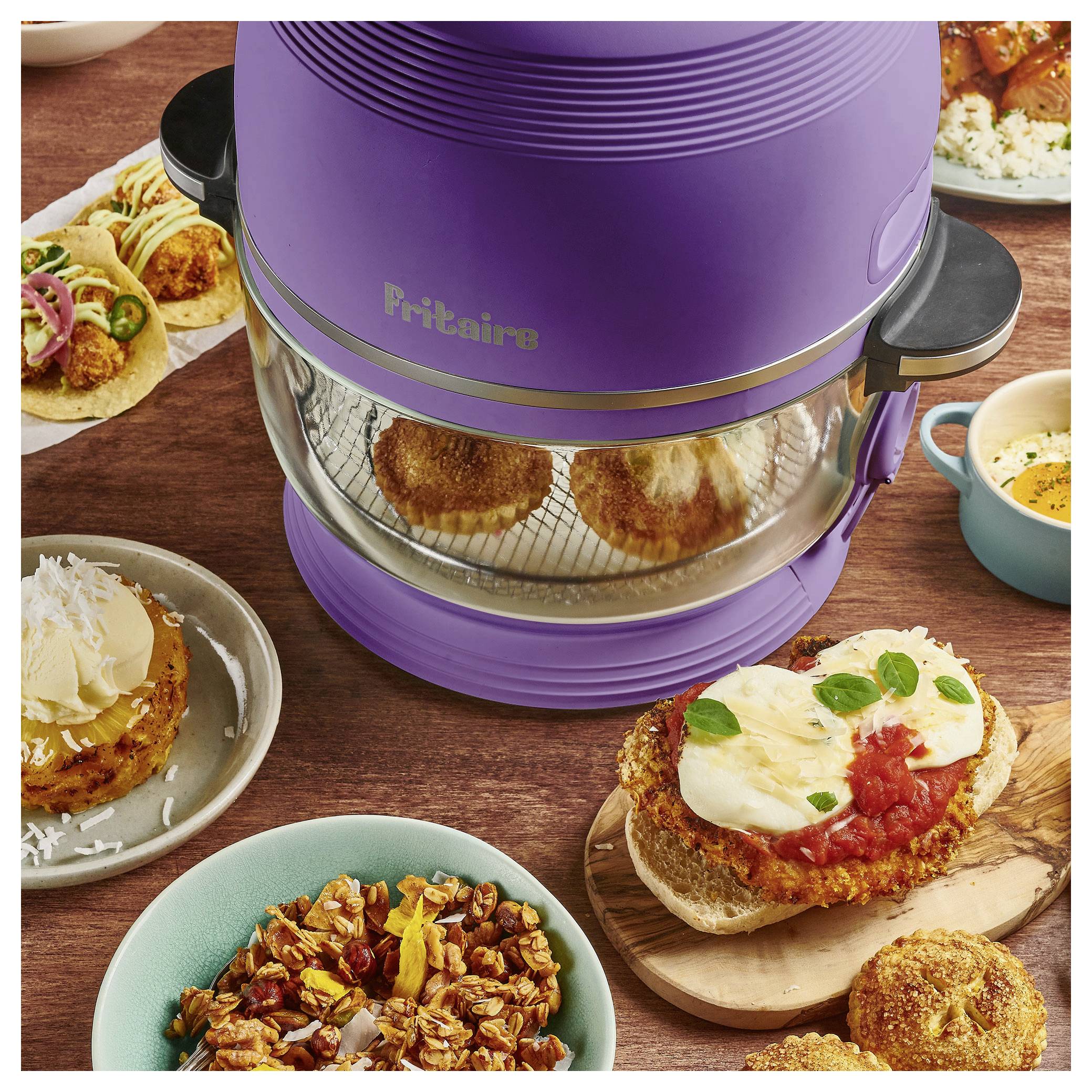 Ein lila Fritaire-Heißluftfritteuse mit zwei fertigen Muffins im Inneren, umgeben von verschiedenen Gerichten auf Tellern, darunter Tacos, Suppe und Gebäck, auf einem Holztisch.