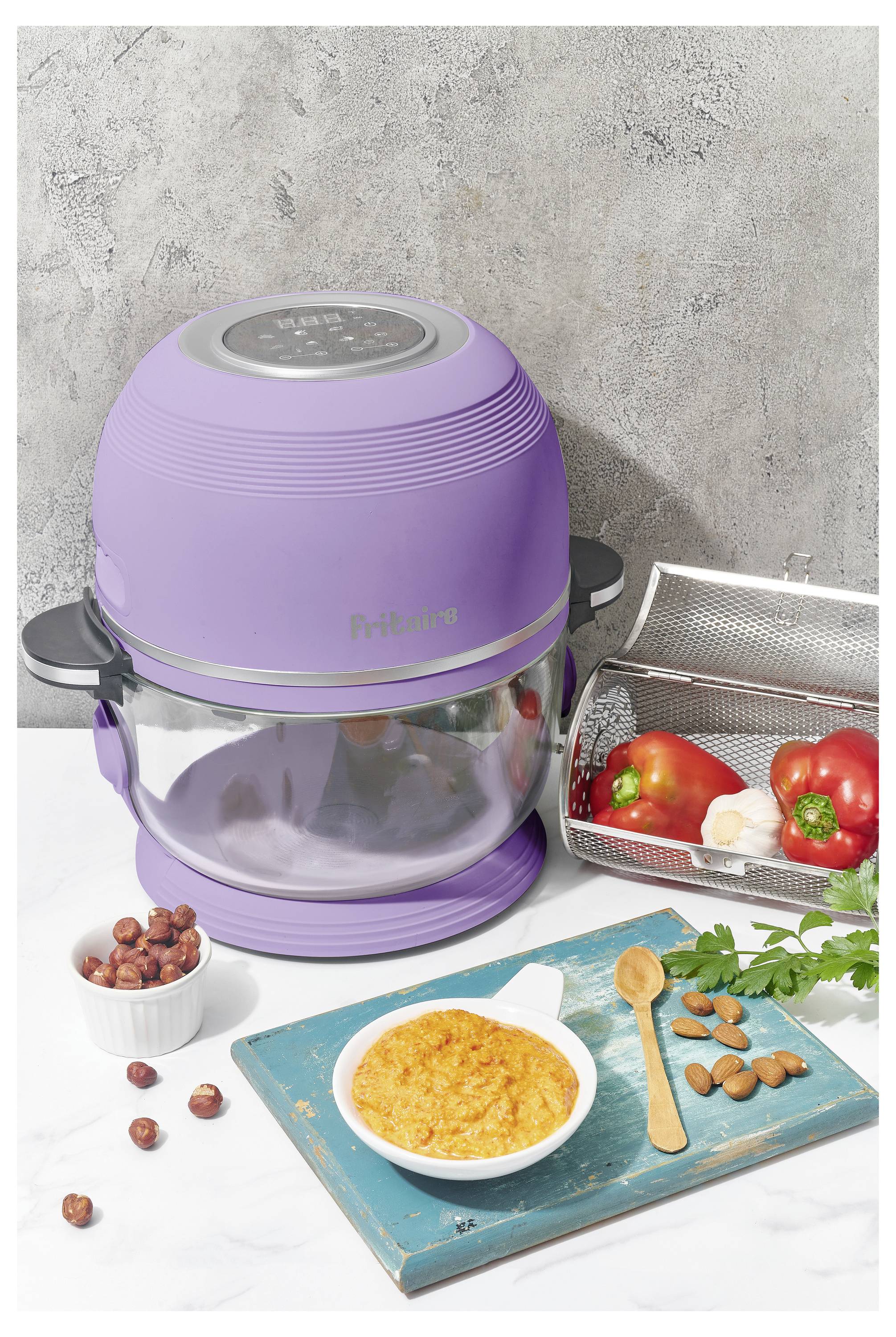 Ein lila Heißluftfritteuse auf einer Küchenarbeitsplatte mit einer Glasschüssel. In der Nähe befinden sich ein Metallkorb mit Gemüse, ein kleines Schälchen mit Nüssen und eine Schüssel mit Suppe.