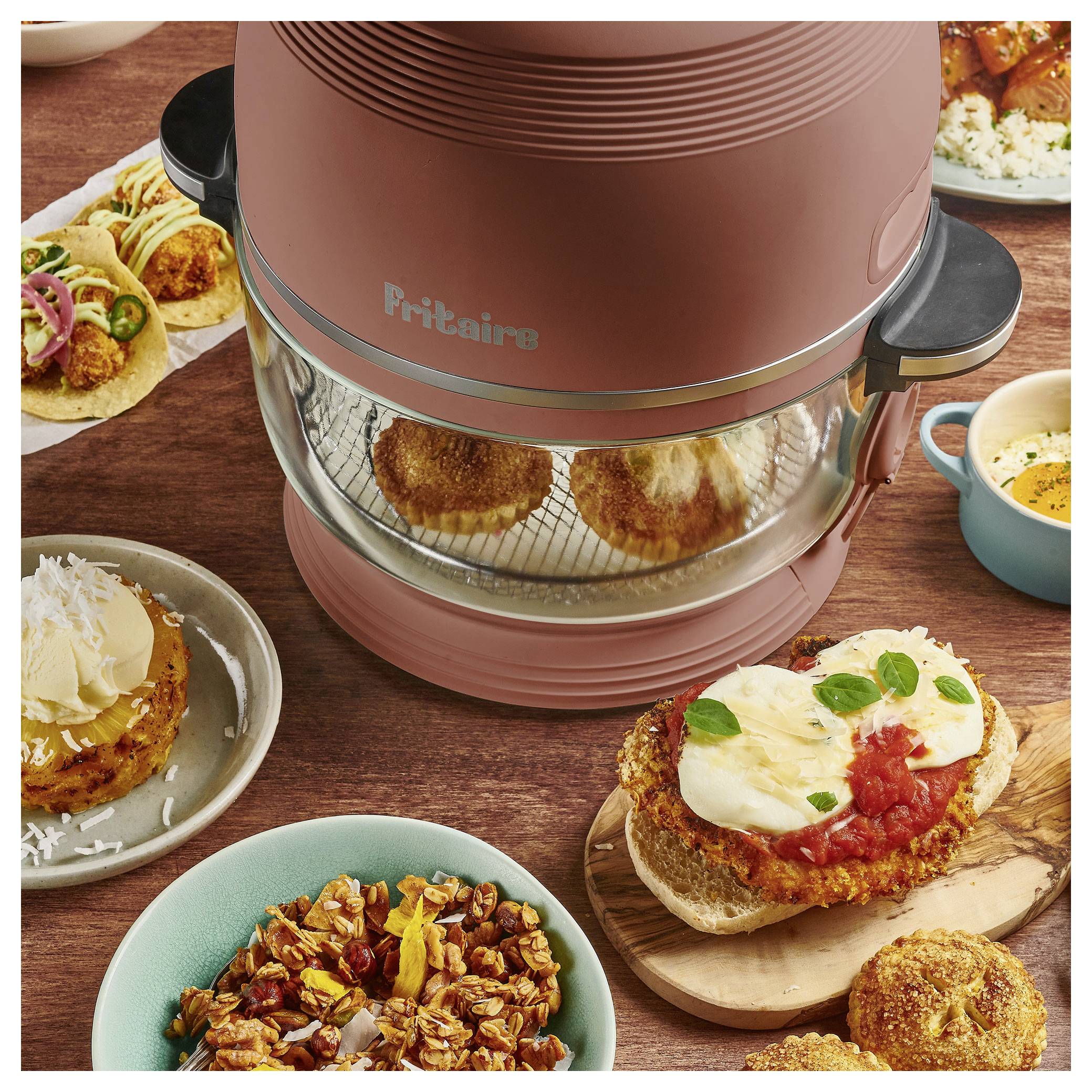 Eine Küchenarbeitsplatte mit einem Heißluftfritteusen, die Lebensmittel enthält. Drumherum sind Teller mit Tacos, ein Burger, der mit Tomatensoße und Käse belegt ist, und eine Schüssel mit Samen.