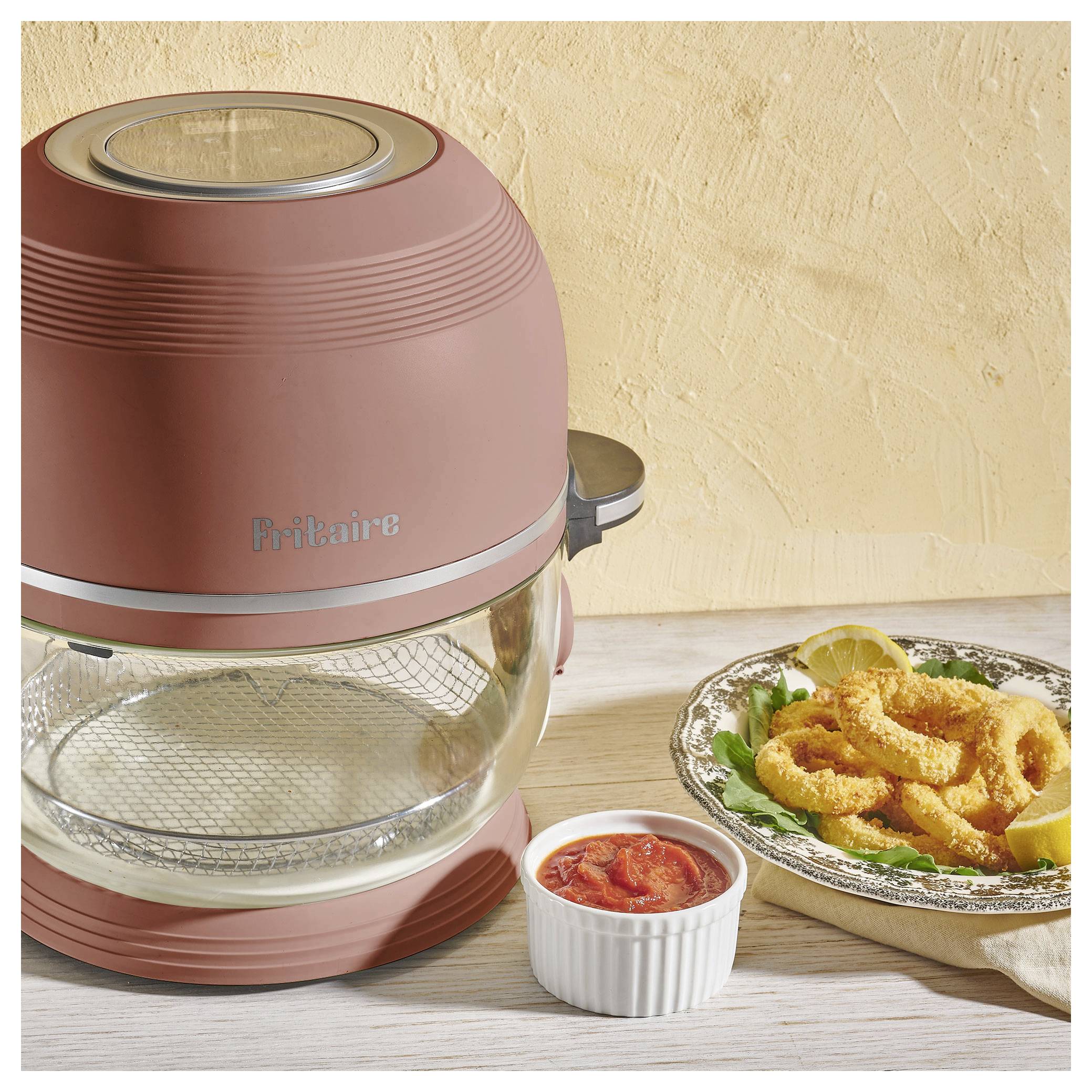 Ein rosa Heißluftfritteuse auf einer Küchenarbeitsplatte mit einer kleinen Schüssel roter Sauce und einem Teller frittierter Speisen, garniert mit Grün und Zitrone.