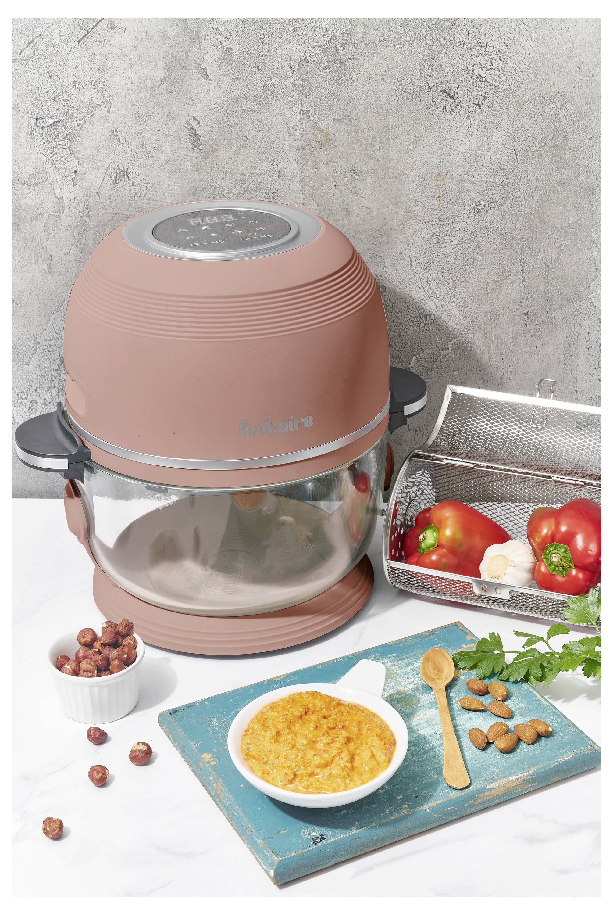 Ein rosa Heißluftfritteuse mit dem Etikett 'Pitalina' neben einem Korb mit Gemüse, einer Schüssel mit Püree und Gewürzen auf einer weiß-türkisfarbenen Oberfläche.