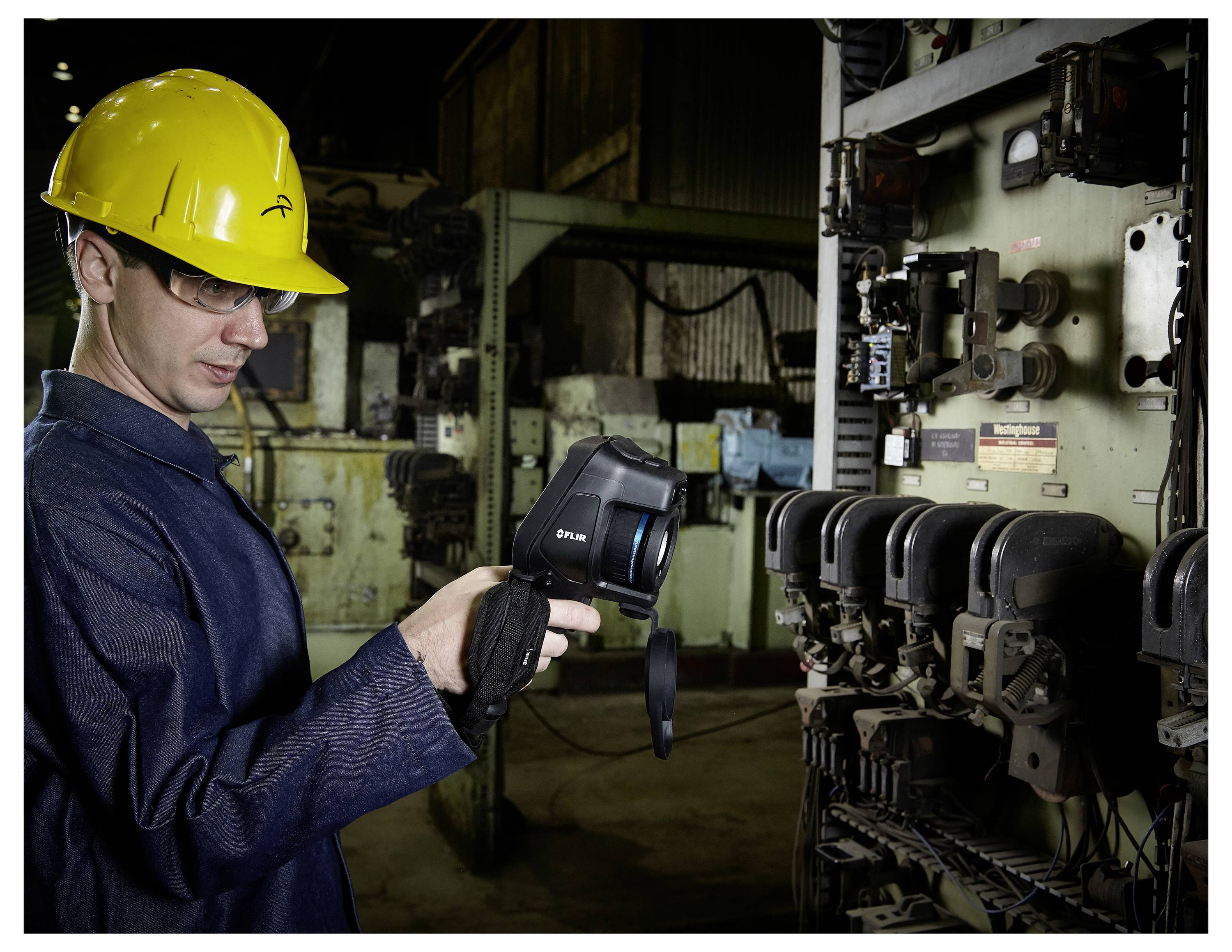 Eine Person mit einem gelben Schutzhelm verwendet eine tragbare Wärmebildkamera zur Inspektion von Industriemaschinen in einer Fabrikumgebung.