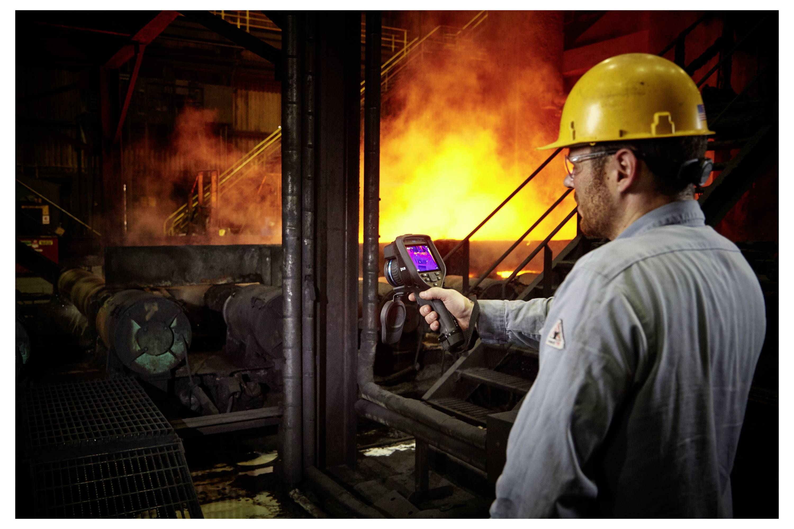 Ein Arbeiter mit Schutzhelm nutzt eine Wärmekamera, um die Temperatur in einer industriellen Umgebung zu überwachen, während im Hintergrund glühendes Metall zu sehen ist.