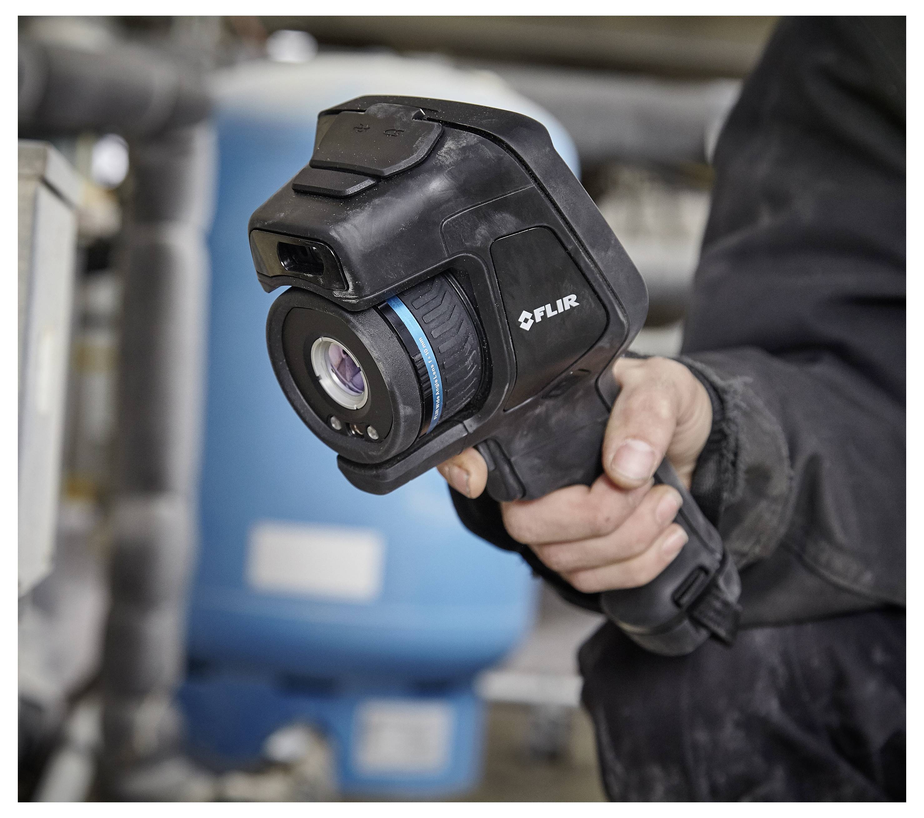 Eine Person hält eine FLIR-Wärmebildkamera, die zur Erkennung von Wärmemustern in einer industriellen Umgebung mit Rohrleitungen und Ausrüstung verwendet wird.