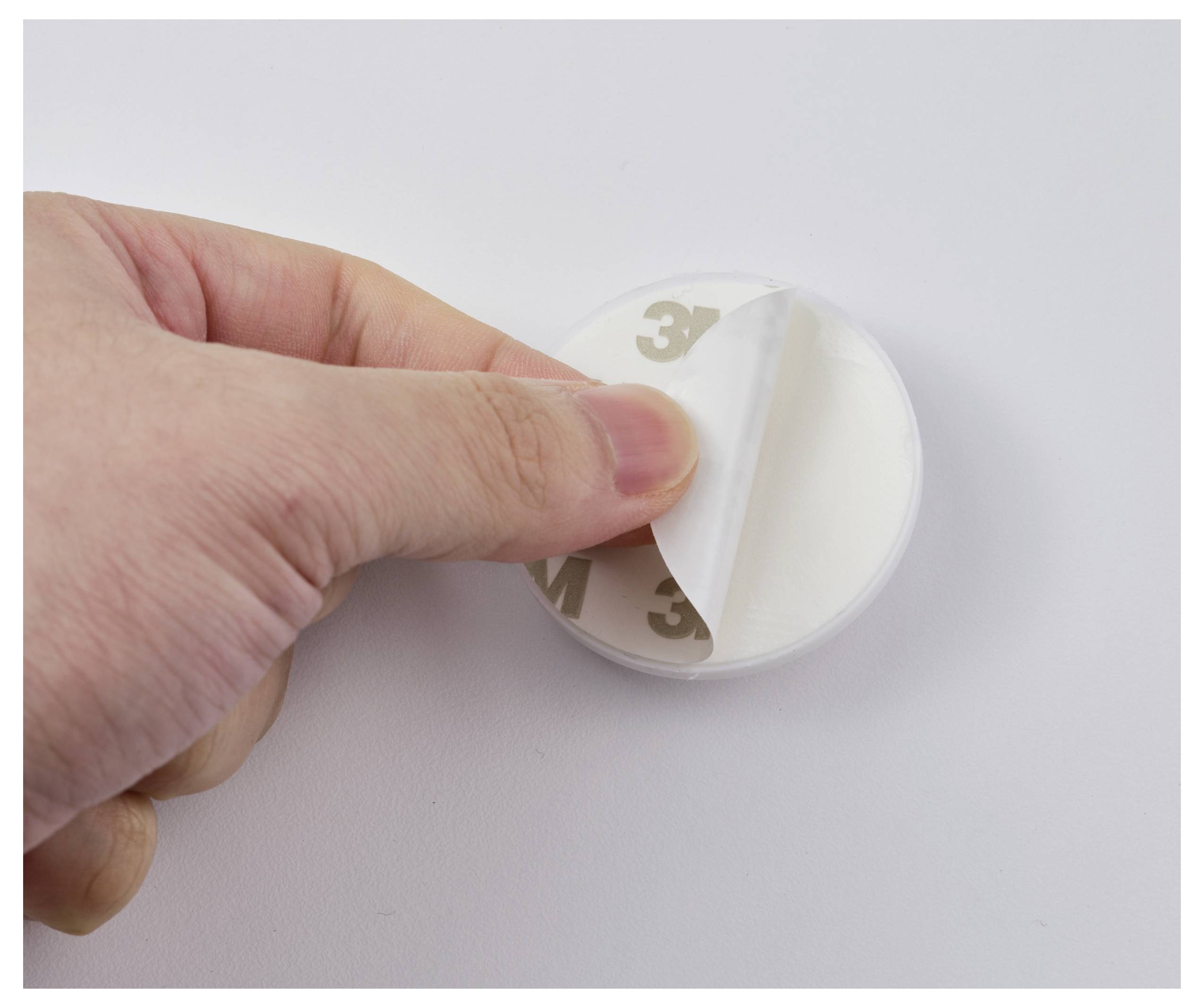 Eine Hand zieht die Rückseite eines runden Klebepads ab und offenbart dessen klebrige Seite zum Anbringen oder Befestigen von Gegenständen.