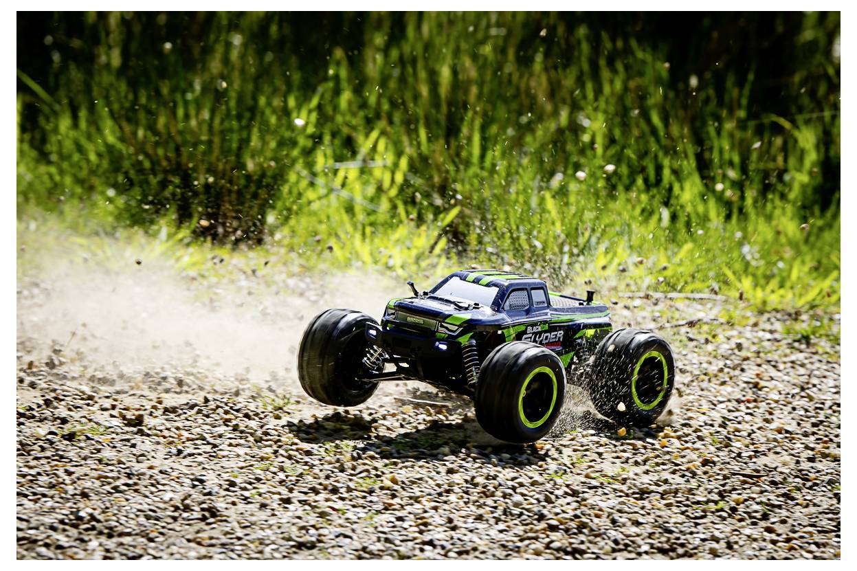Ein ferngesteuertes Truck mit großen Reifen wirbelt Staub auf, während es über einen Kiesweg rast, umgeben von grünem Gras.