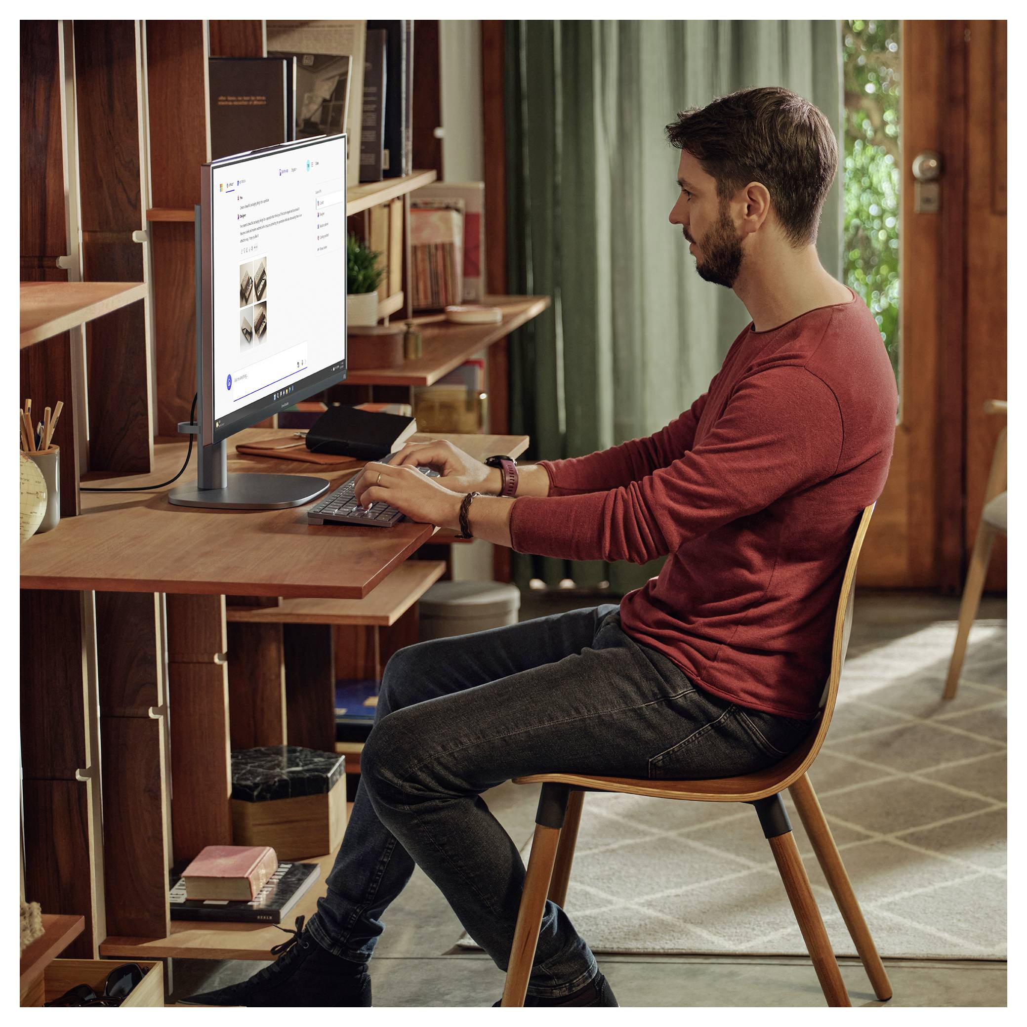 Eine Person in einem roten Hemd sitzt an einem Schreibtisch und arbeitet an einem Computer. Der Raum verfügt über Holzregale, einen geschlossenen Vorhang und verschiedene persönliche Gegenstände.