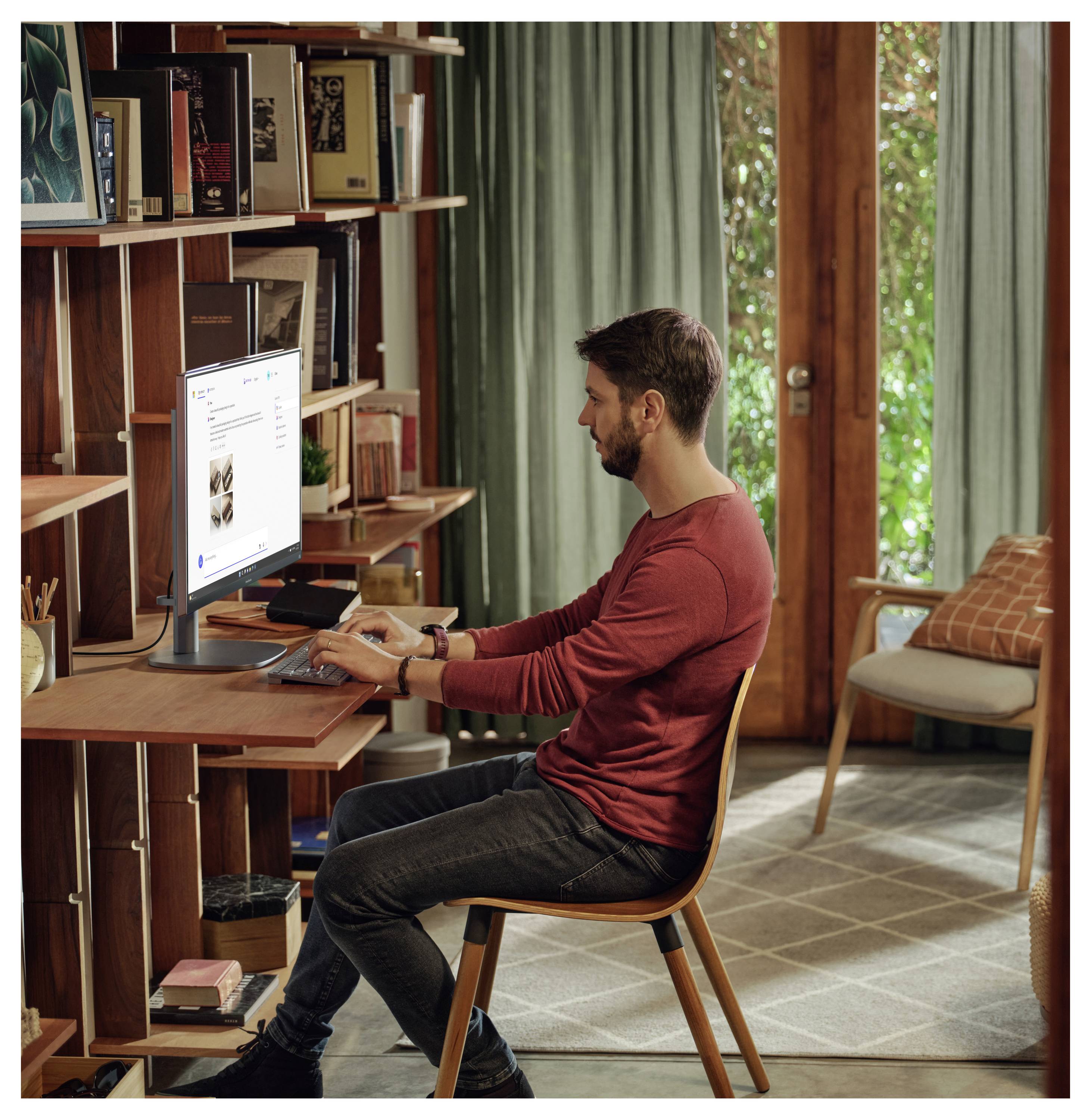 Ein Mann in einem roten Pullover sitzt an einem Holzschreibtisch und blickt auf einen Computerbildschirm in einem gemütlichen Raum mit Bücherregalen und großen Fenstern.