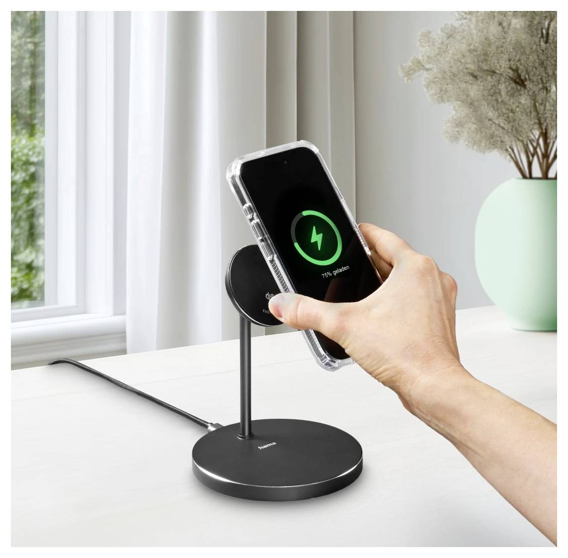 Eine Hand legt ein Smartphone auf einen schwarzen drahtlosen Ladegerät, wobei ein Ladesymbol angezeigt wird. Im Hintergrund steht eine Vase mit getrockneten Blumen.