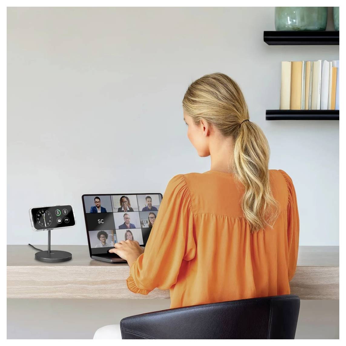 Eine Frau in einer orangefarbenen Bluse führt eine Videokonferenz auf einem Tablet mit mehreren Personen. Ein Smart-Home-Gerät steht auf dem Tisch. Bücher sind in Regalen aufgestellt.