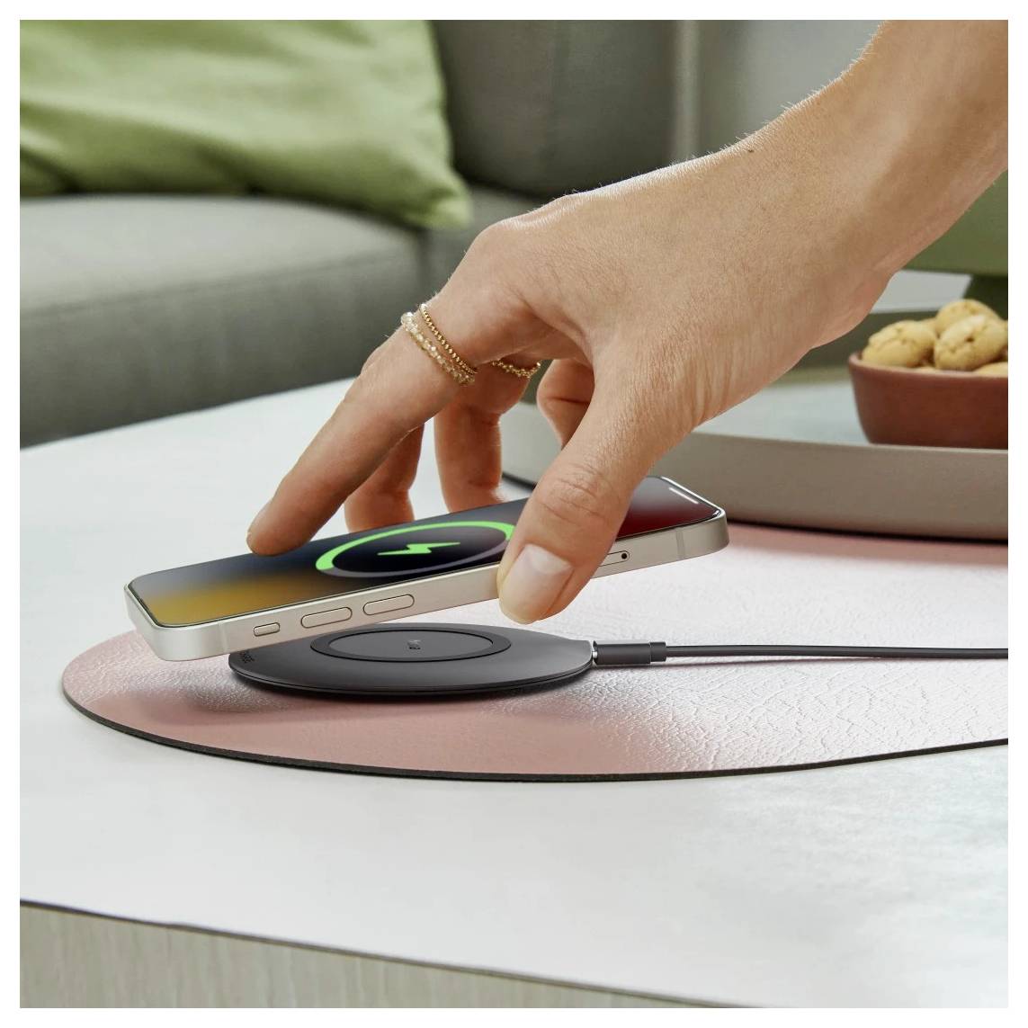 Eine Hand, die ein Smartphone auf ein kabelloses Ladegerät legt, wobei ein Ladesymbol auf dem Bildschirm sichtbar ist, auf einem Tisch in einer gemütlichen Inneneinrichtung.