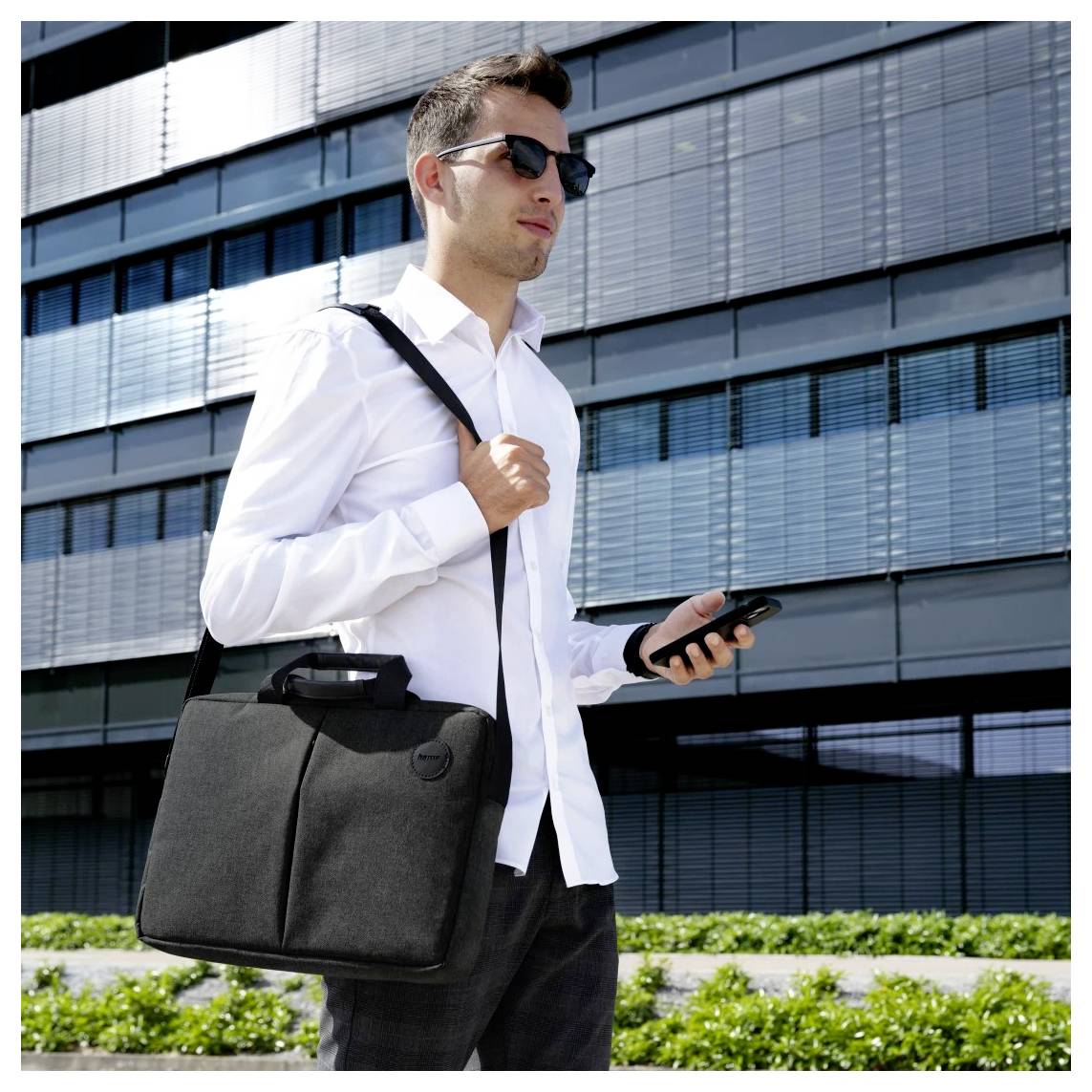 Ein Mann in einem weißen Hemd und Sonnenbrille hält ein Telefon und einen schwarzen Aktenkoffer und steht vor einem modernen Glasgebäude.