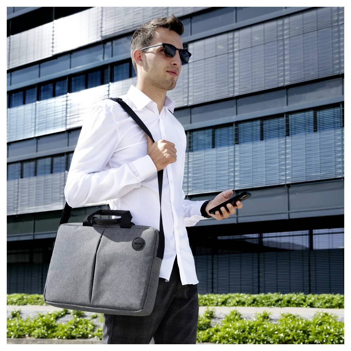 Eine Person in Geschäftskleidung geht vor einem modernen Bürogebäude nach draußen, hält einen Aktenkoffer und prüft ihr Smartphone.