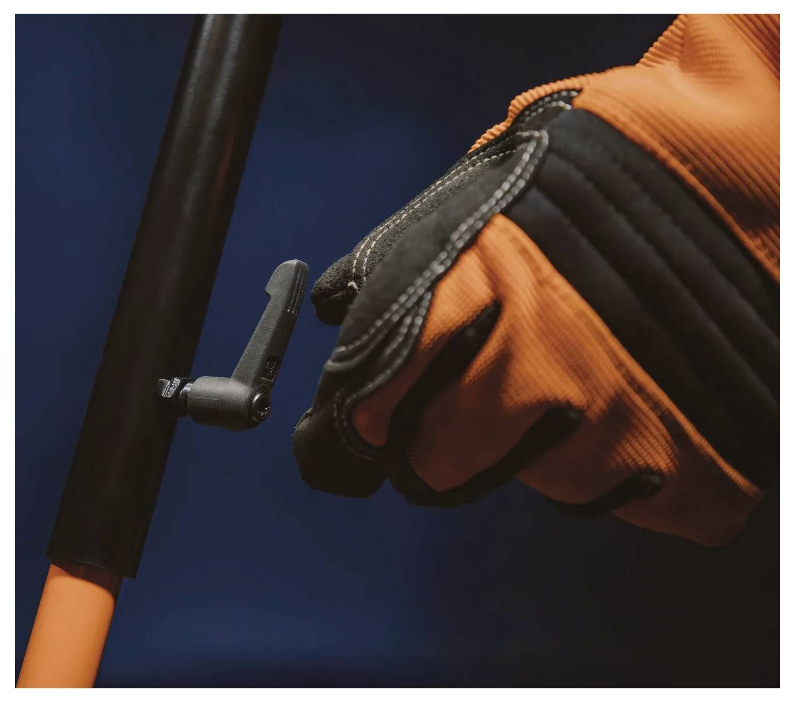 Eine Hand mit Handschuh umfasst einen Hebel an einem Holm vor einem dunklen Hintergrund und deutet auf die Steuerung oder Bedienung eines Werkzeugs oder Geräts hin.