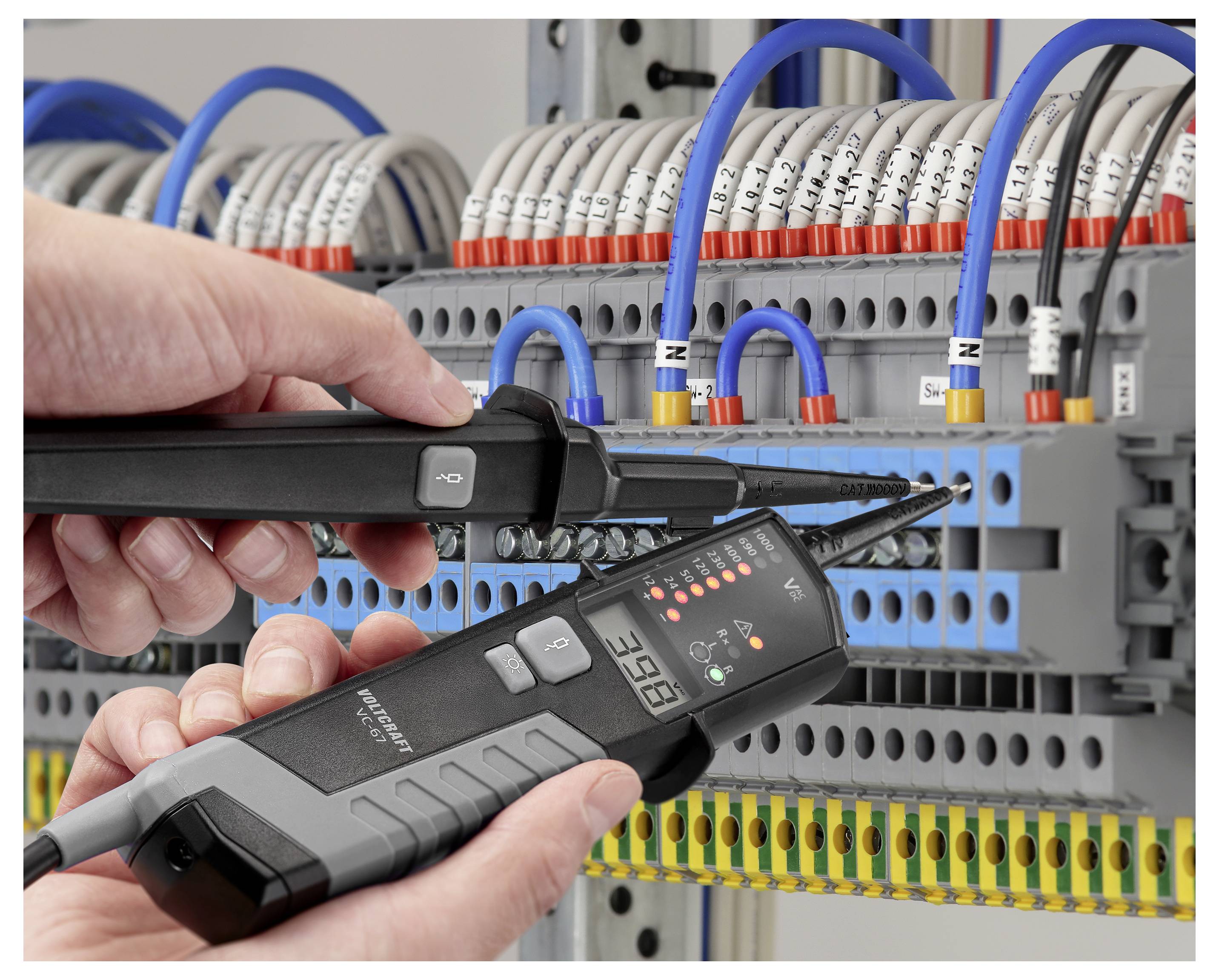 Hände testen ein elektrisches Steuerpanel mit zwei Spannungs-/Strommessgeräten zwischen Klemmleisten mit Verdrahtung und Schaltverbindungen.