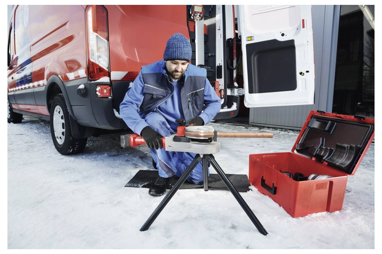Eine Person in Arbeitskleidung bedient ein Rohwerkzeug im Freien neben einem roten Transporter mit geöffneten Hecktüren. Eine rote Werkzeugkiste befindet sich in der Nähe auf dem verschneiten Boden.