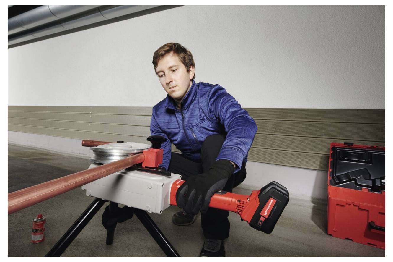 Eine Person in einer blauen Jacke verwendet ein Elektrowerkzeug, um ein Metallrohr zu biegen. Sie kniet neben einem Werkzeugkasten auf einem Betonboden.