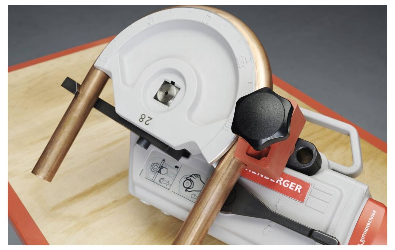 Ein Rohrbieger aus Kupfer auf einer Holzoberfläche mit einem teilweise gebogenen Rohr, der den Biegeprozess und den Mechanismus des Werkzeugs detailliert zeigt.