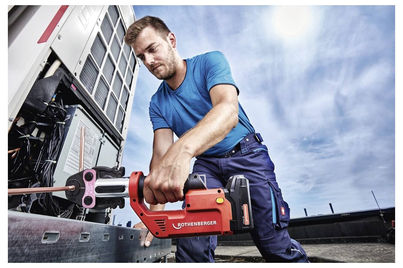 Ein Techniker in blauer Arbeitskleidung bedient ein rotes hydraulisches Werkzeug und arbeitet konzentriert an einer großen Maschine im Freien unter einem bewölkten Himmel.