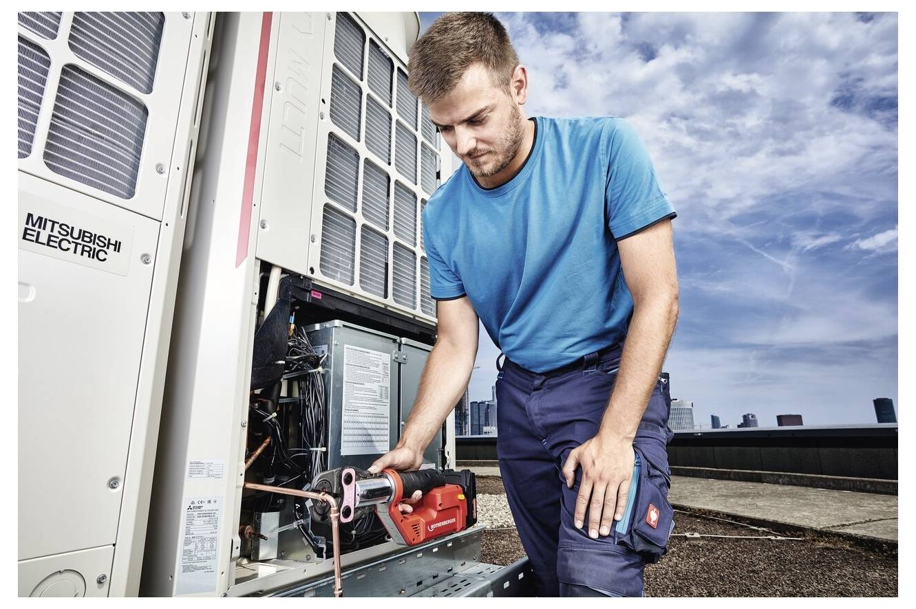 Ein Techniker in einem blauen Hemd und Arbeitskleidung repariert eine industrielle Klimaanlage mit einem Werkzeug auf einem Dach, mit einer Stadtsilhouette im Hintergrund.