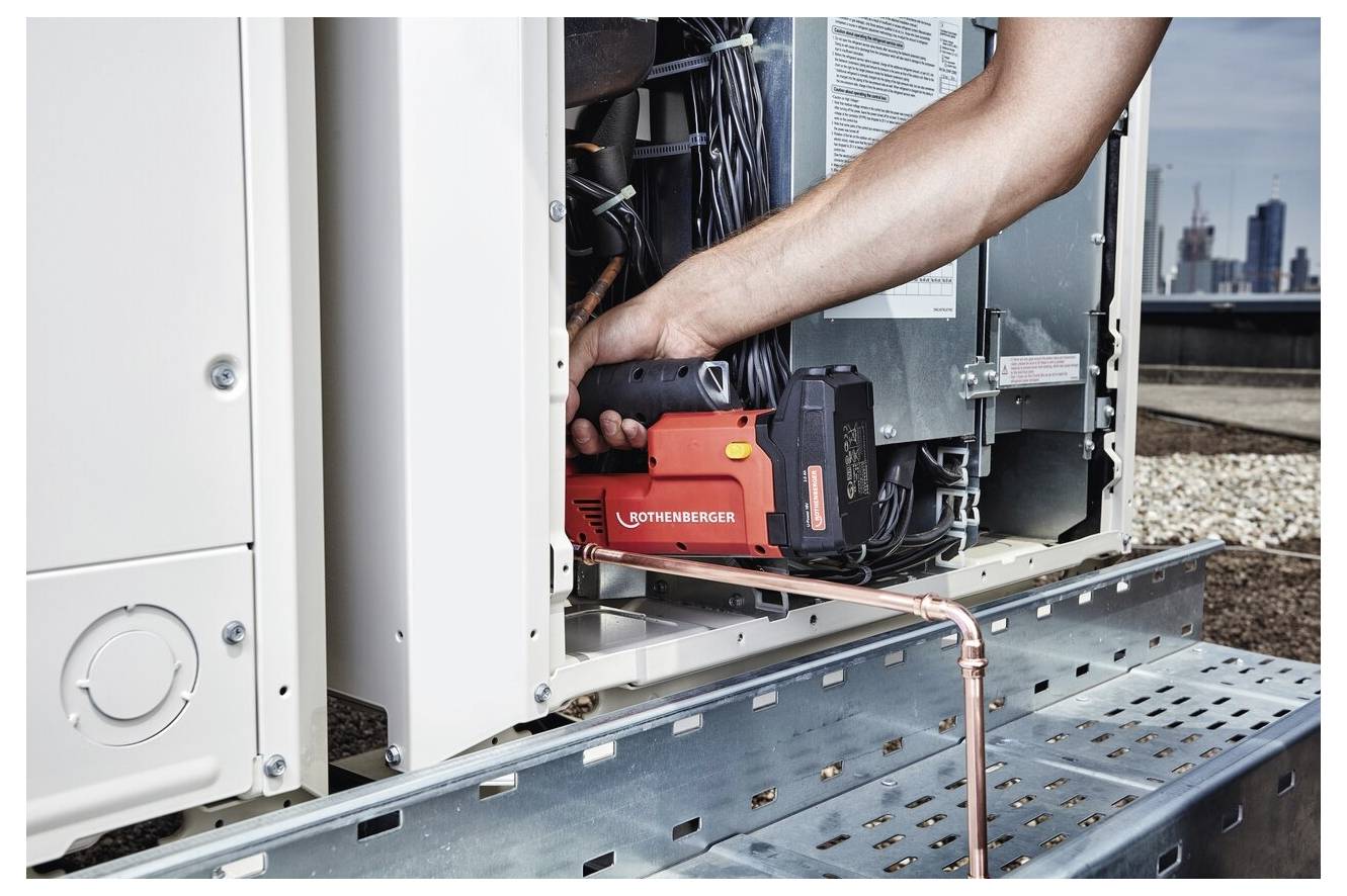 Eine Person, die ein rotes Rothenberger-Werkzeug bei der Arbeit an einer elektrischen Einheit auf einem Dach verwendet, wobei Stadtgebäude im Hintergrund sichtbar sind.