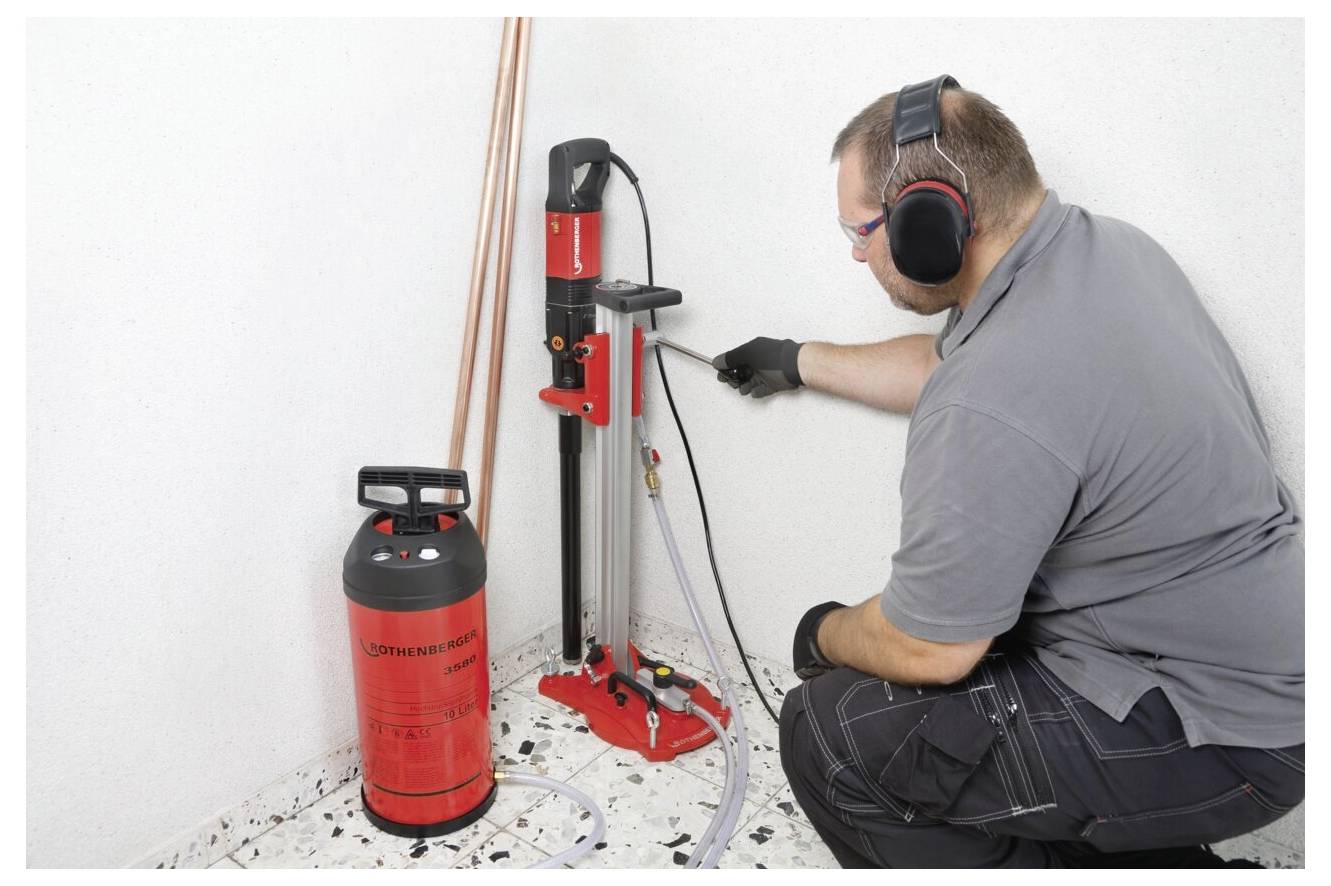 Eine Person mit Ohrenschützern bedient eine rote Wandbohrmaschine in der Nähe einer weißen Wand, wobei ein zylindrischer Gaskontainer neben ihr auf dem Boden steht.