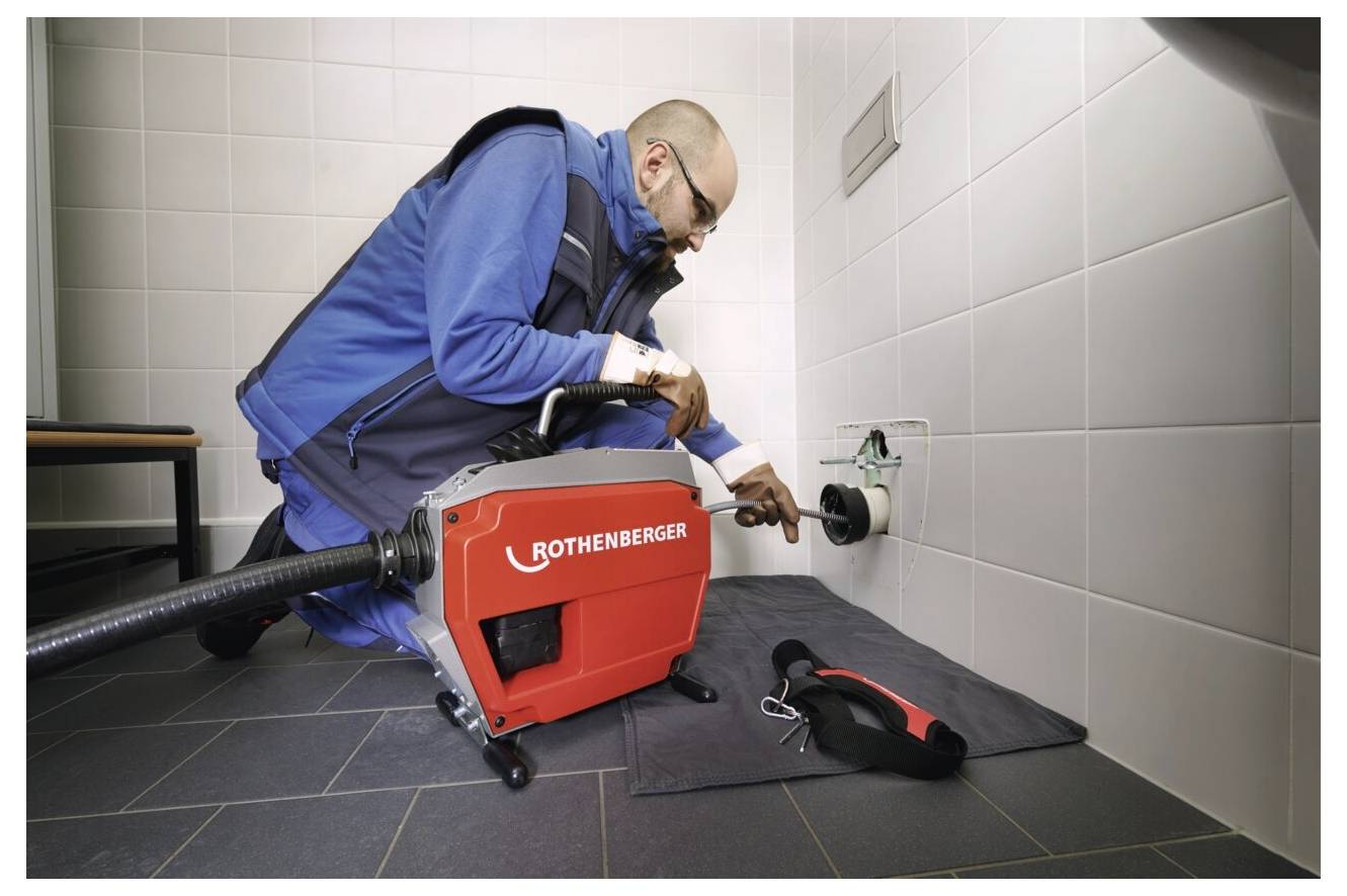 Eine Person in Arbeitskleidung verwendet ein Rothenberger-Werkzeug, um eine Wandrohrleitung in einem gefliesten Raum zu warten. Werkzeuge sind auf einer Matte in der Nähe platziert.