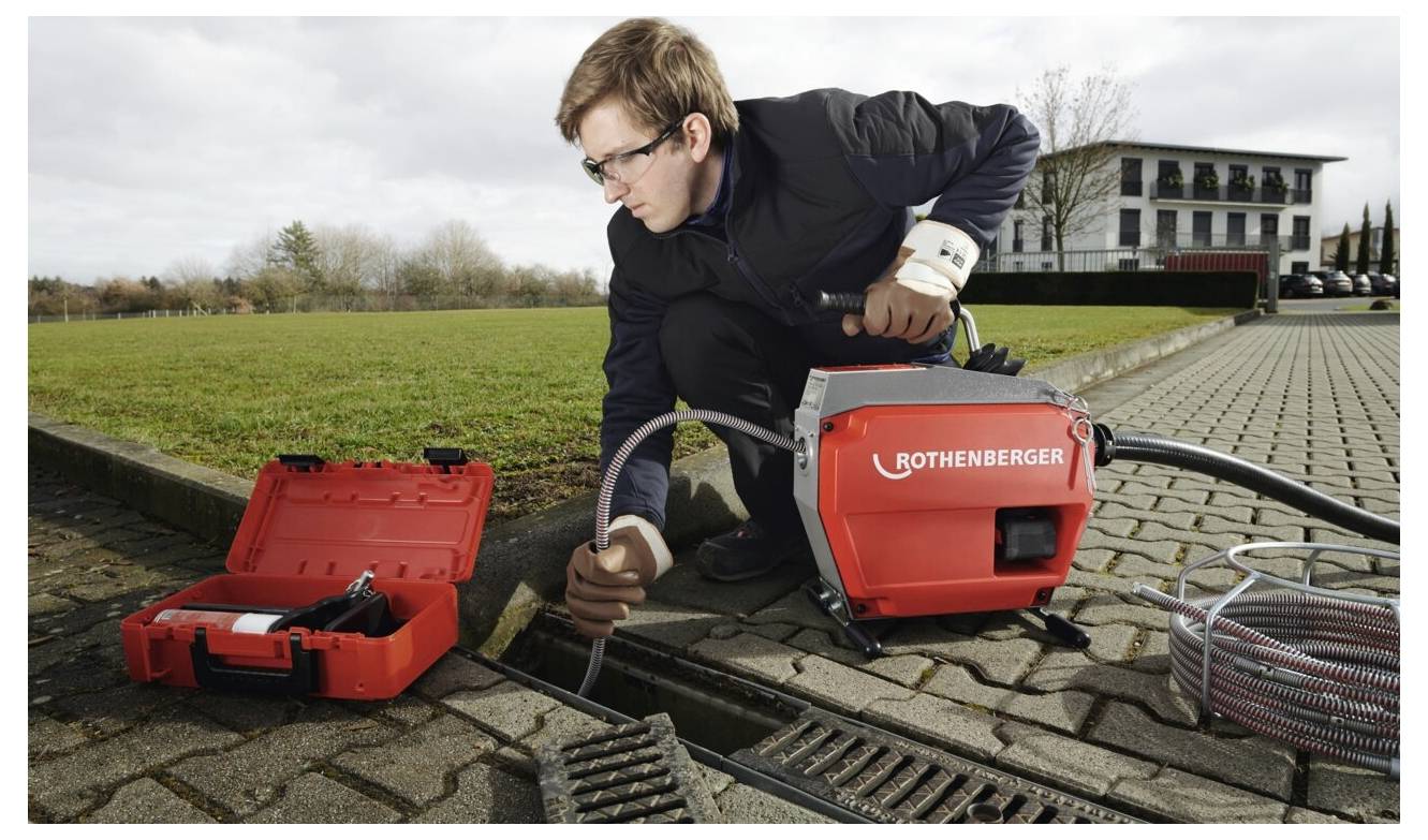 Eine Person benutzt eine Rothenberger Rohrreinigungsmaschine, kniet neben einem Außenablauf mit einer geöffneten Werkzeugkiste in der Nähe, unweit eines Gebäudes.