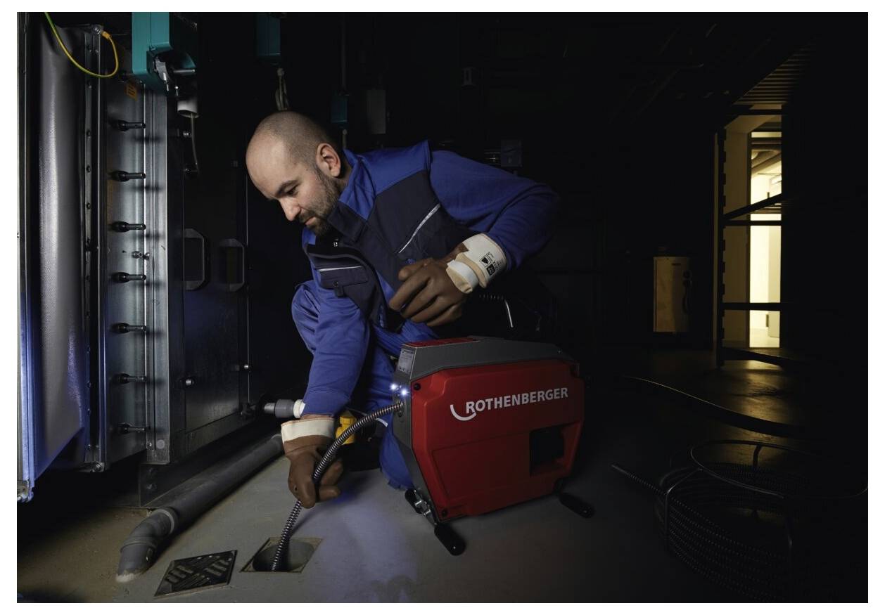 Eine Person, die eine Rothenberger-Maschine zum Reinigen eines Abflussrohrs verwendet, trägt eine blaue Arbeitsuniform und befindet sich in einer düsteren industriellen Umgebung.
