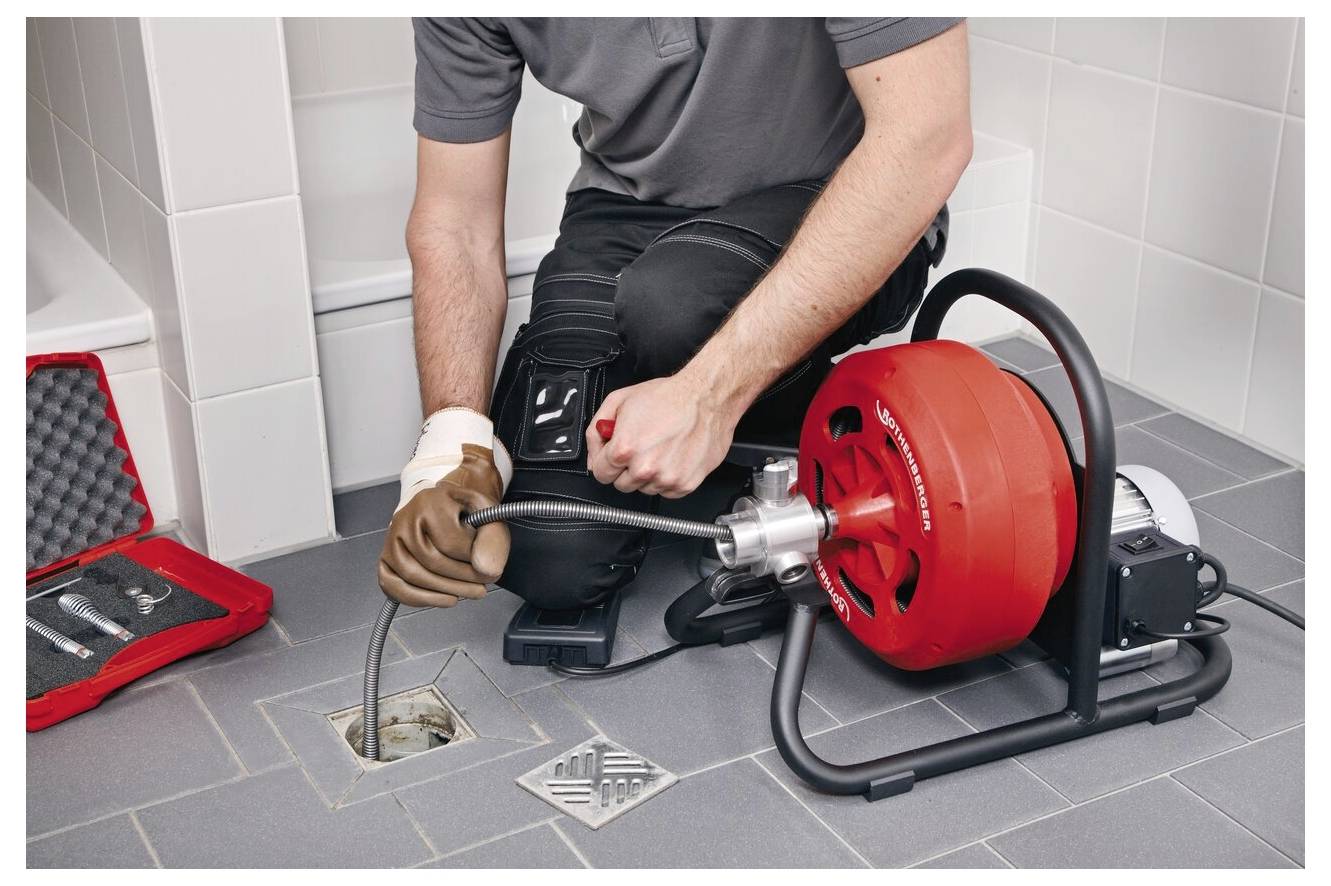 Ein Klempner benutzt einen mechanischen Rohrreiniger, um einen Abfluss im Badezimmerboden zu entfernen. Ein Werkzeugset ist in der Nähe geöffnet und zeigt verschiedene Werkzeuge.