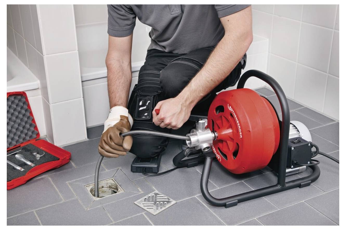 Eine Person benutzt eine rote Rohrreinigungsmaschine, um einen Bodenablauf in einem gefliesten Badezimmer zu entstopfen. Eine Werkzeugausrüstung liegt in der Nähe geöffnet.