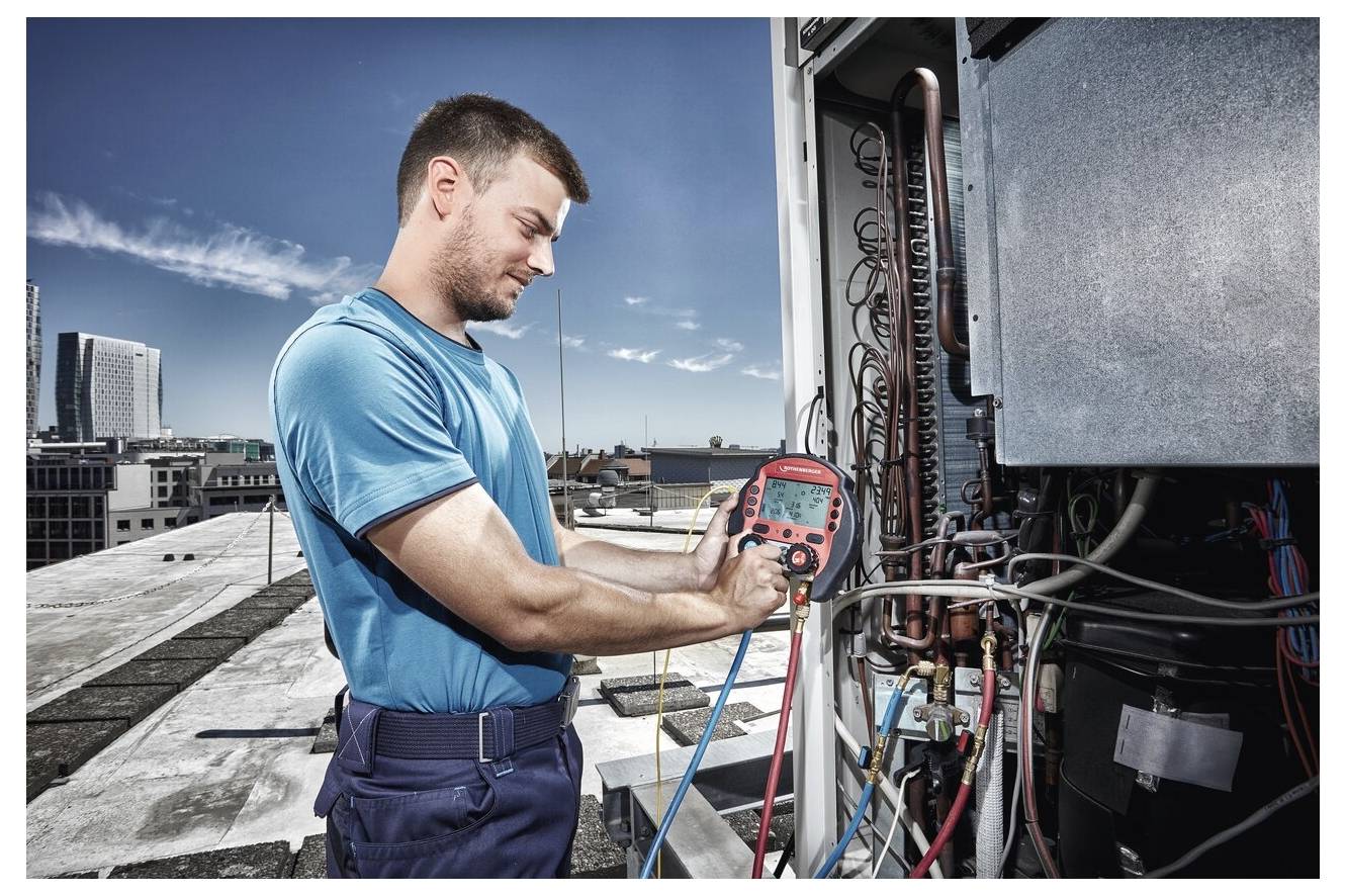 Ein Techniker in einem blauen Hemd verwendet ein Multimeter, um Klimaanlagen auf einem Dach zu überprüfen, mit einer Stadtkulisse im Hintergrund.