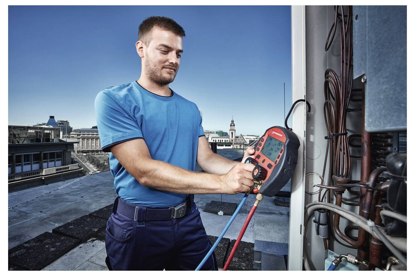 Ein Techniker in einem blauen Hemd bedient Diagnosegeräte an einer Dacheinheit einer Klimaanlage, mit einer Stadtskyline im Hintergrund.