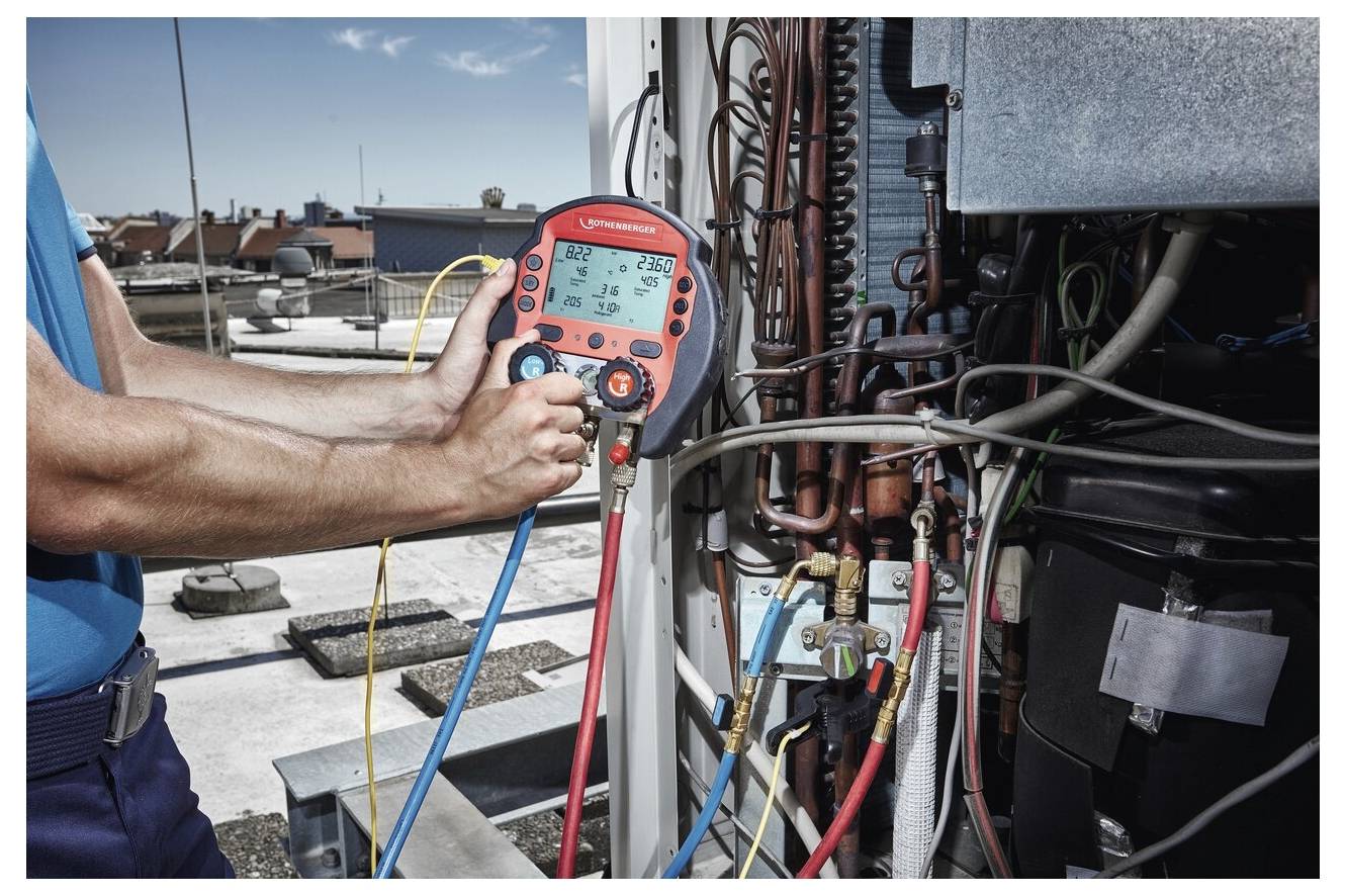 Ein Techniker benutzt einen digitalen Druckmesser an einer HVAC-Anlage und überprüft die Kältemittelstände. Im Hintergrund sind Dachaufbauten und ein klarer Himmel zu sehen.