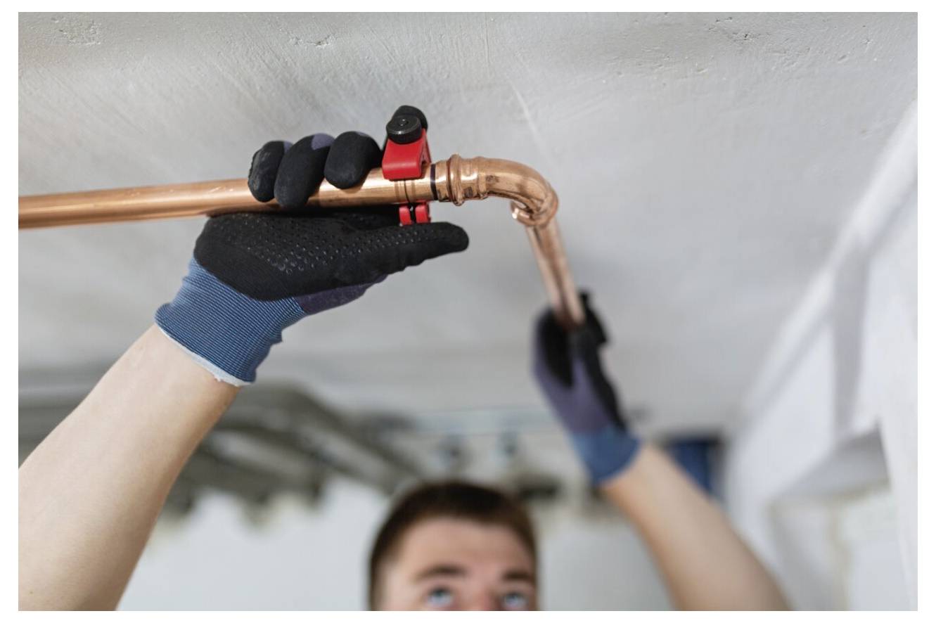 Eine Person mit Handschuhen benutzt ein Werkzeug, um ein Kupferrohr unter einer Decke zu befestigen, was auf Sanitär- oder Heimreparaturarbeiten hinweist.