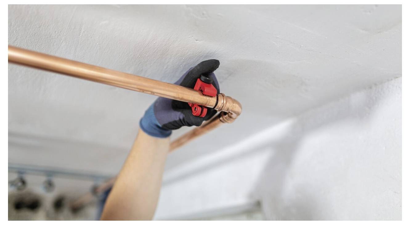 Eine Person mit Handschuhen verwendet einen Rohrabschneider, um ein Kupferrohr zu durchtrennen, das an einer Decke befestigt ist, was auf Sanitär- oder Renovierungsarbeiten hindeutet.