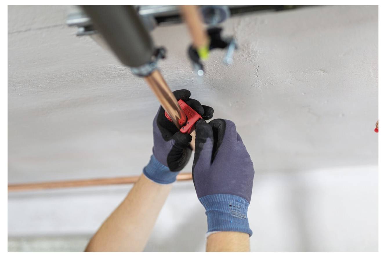 Hände in Handschuhen, die mit einem Schraubenschlüssel ein Kupferrohr an der Decke befestigen.