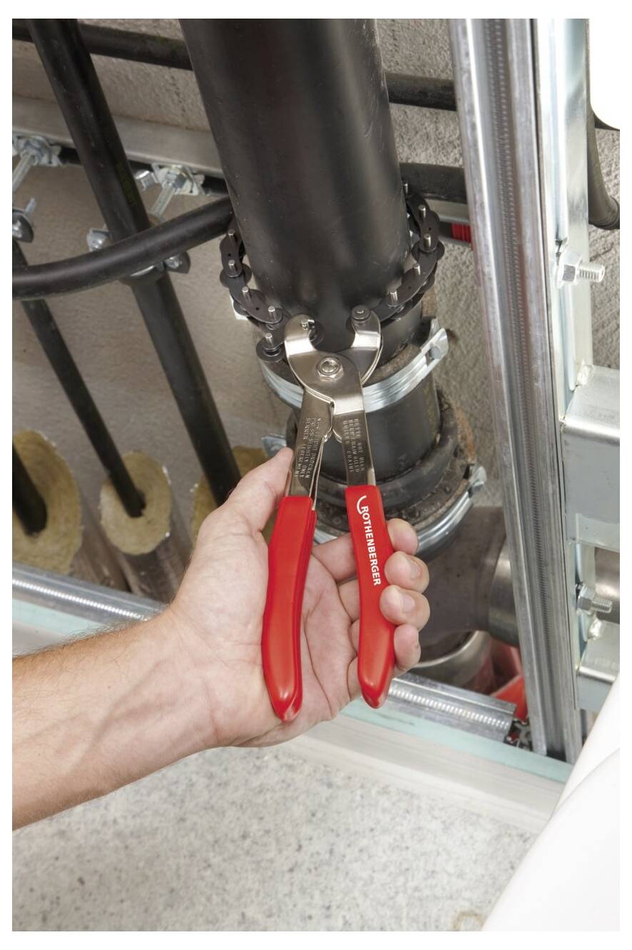 Eine Hand benutzt Zangen mit roten Griffen, um einen Bolzen an einem großen schwarzen Rohr innerhalb eines Metallrahmens zu befestigen, was Klempner- oder Wartungsarbeiten andeutet.