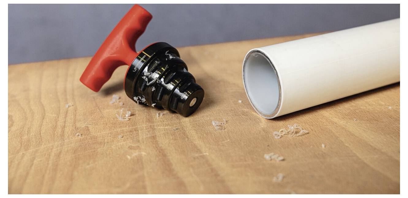 Ein Rohrabschneider mit rotem Griff liegt auf einer Holzoberfläche neben einem weißen PVC-Rohr. Späne sind verstreut, was auf eine kürzliche Schneidaktivität hindeutet.