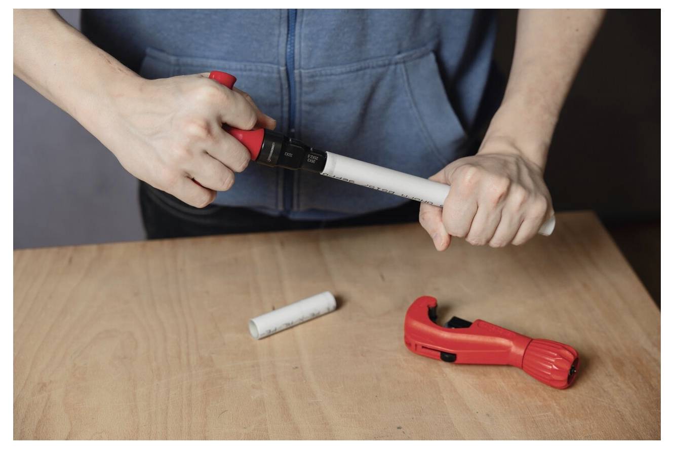 Eine Person verwendet einen roten Rohrabschneider, um ein weißes Plastikrohr auf einem Tisch zu durchtrennen, wobei sich ein weiteres Rohrstück und ein Werkzeug in der Nähe befinden.